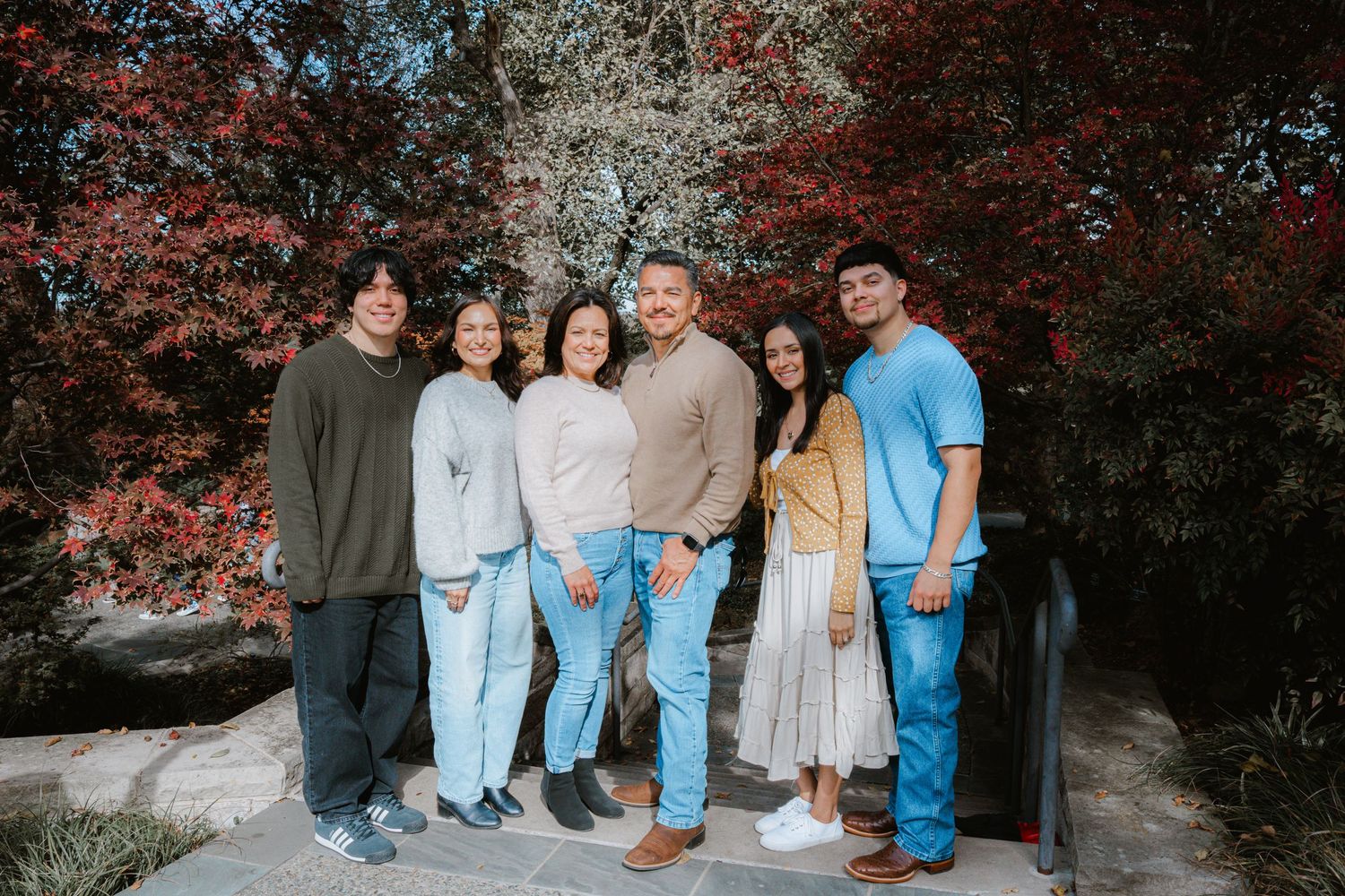 A group stands together outdoors against a backdrop of red autumn foliage and stone walls.