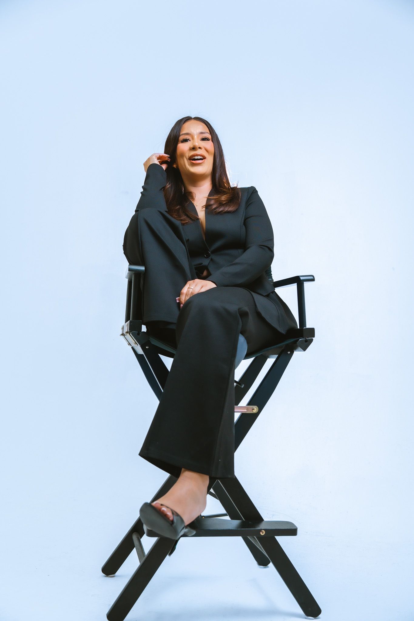 Someone in a black business suit laughs joyfully while sitting in a tall director's chair against a blue background.