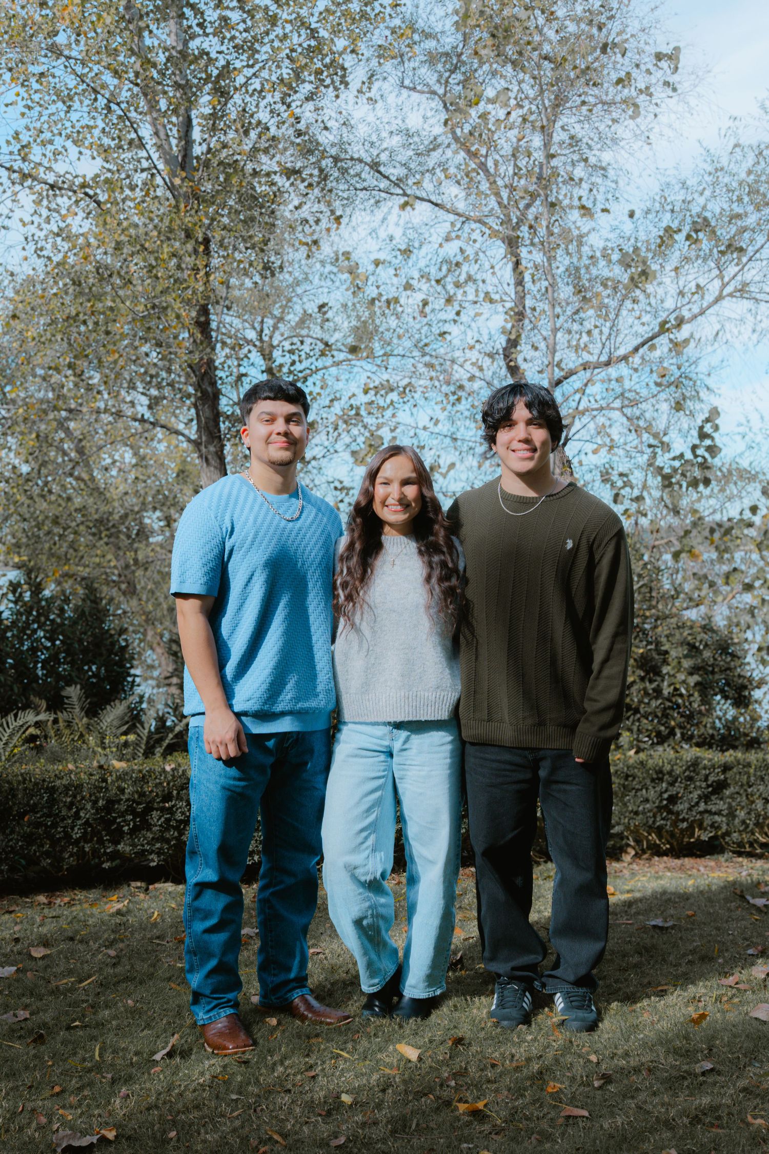 Three people stand together on a grassy hillside in casual clothing on an autumn day.