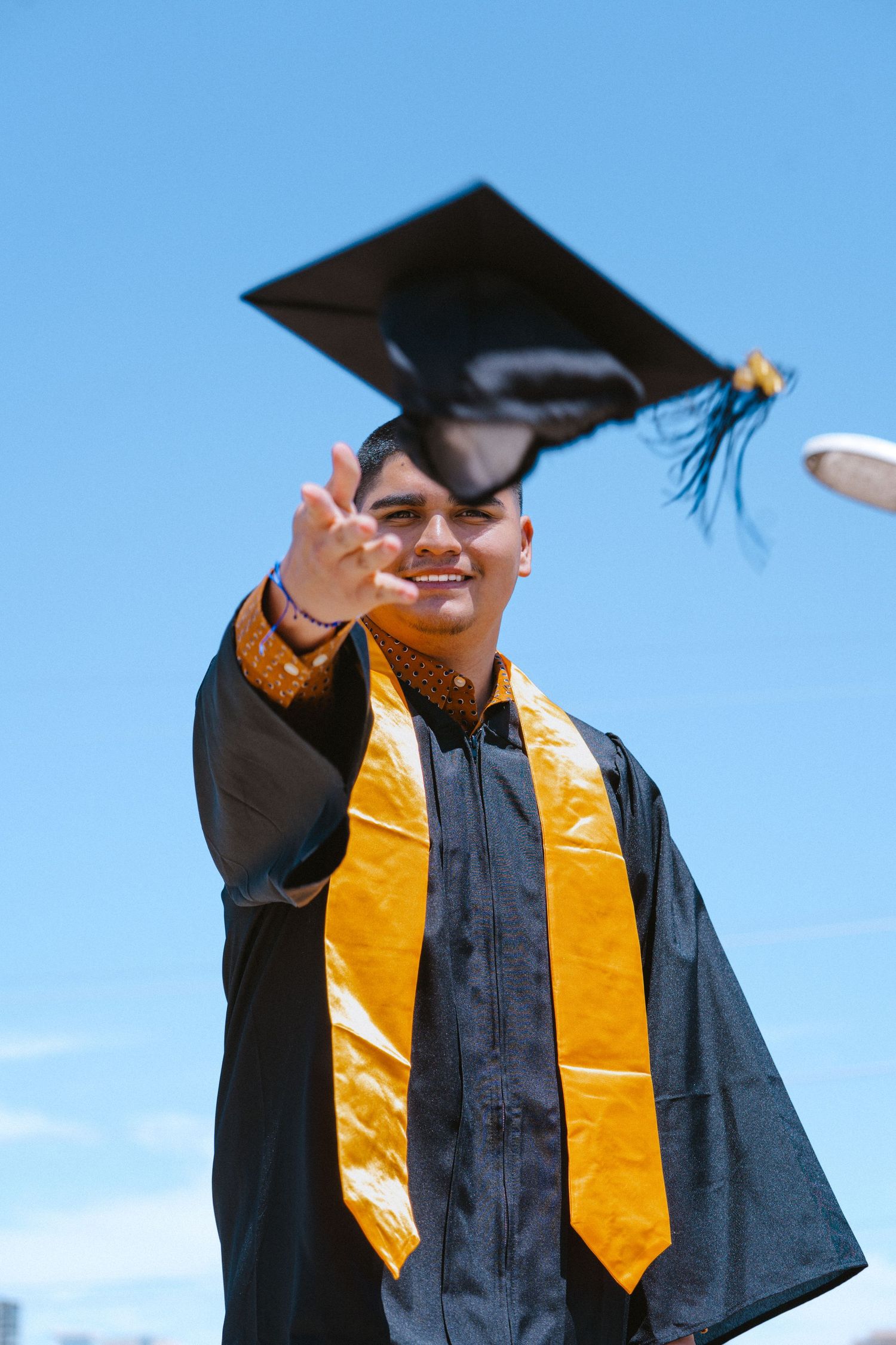 Smiling graduate in black cap and gown with gold stole tossing mortarboard against blue sky.