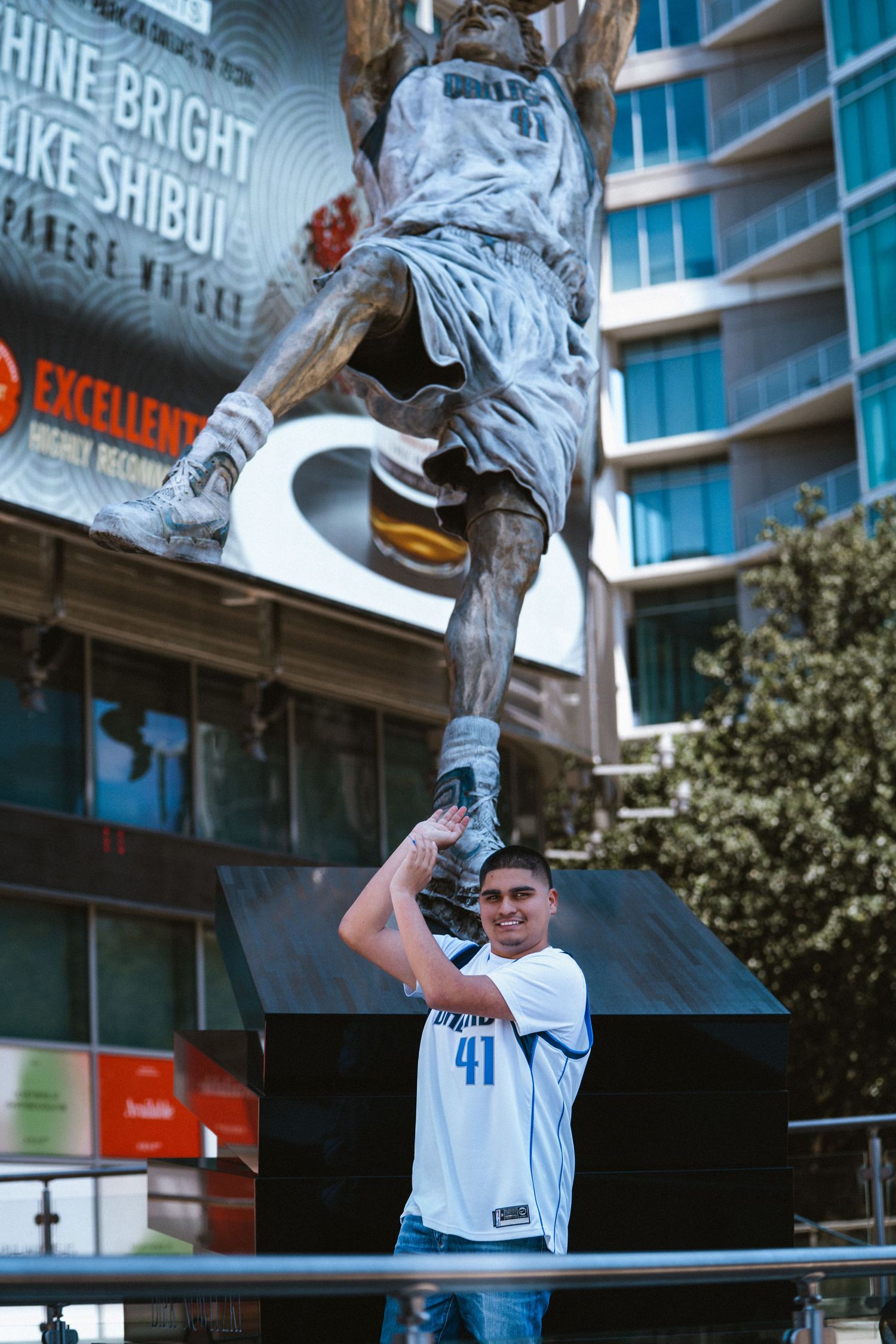 Man in Dirk Nowitzki #41 Mavericks jersey poses in front of Dirk Nowitzki statue outside American Airlines Center in Dallas.