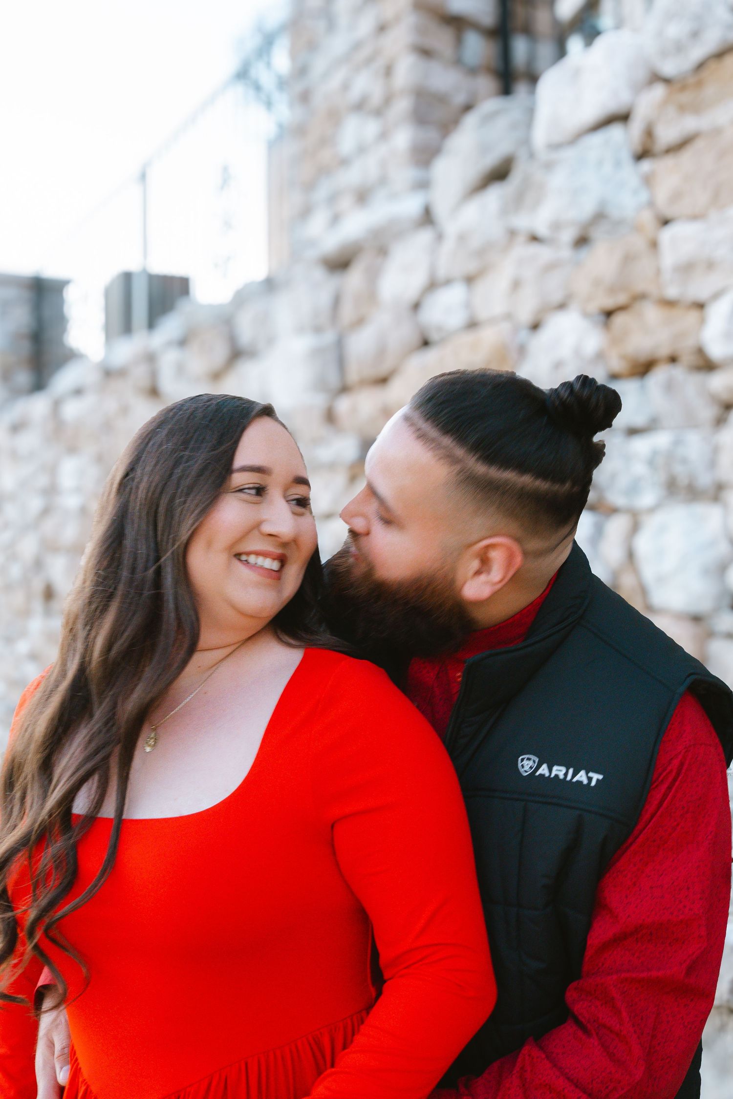 Couple in matching red outfits share a romantic moment against a stone wall, man kissing woman's cheek.