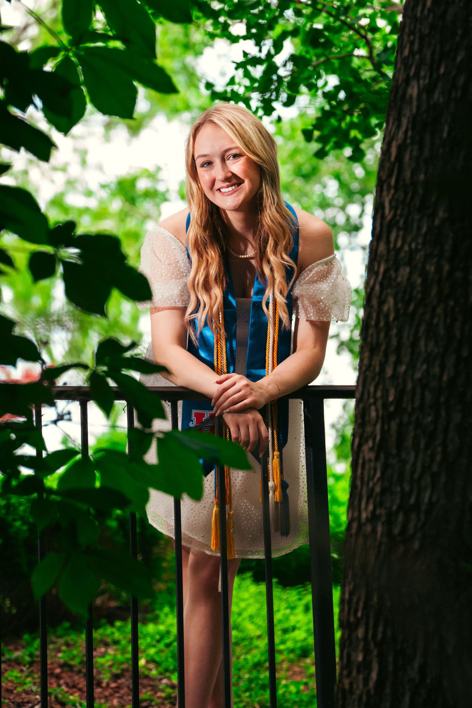 A person in casual attire leans on a metal railing surrounded by lush green foliage in a park setting.