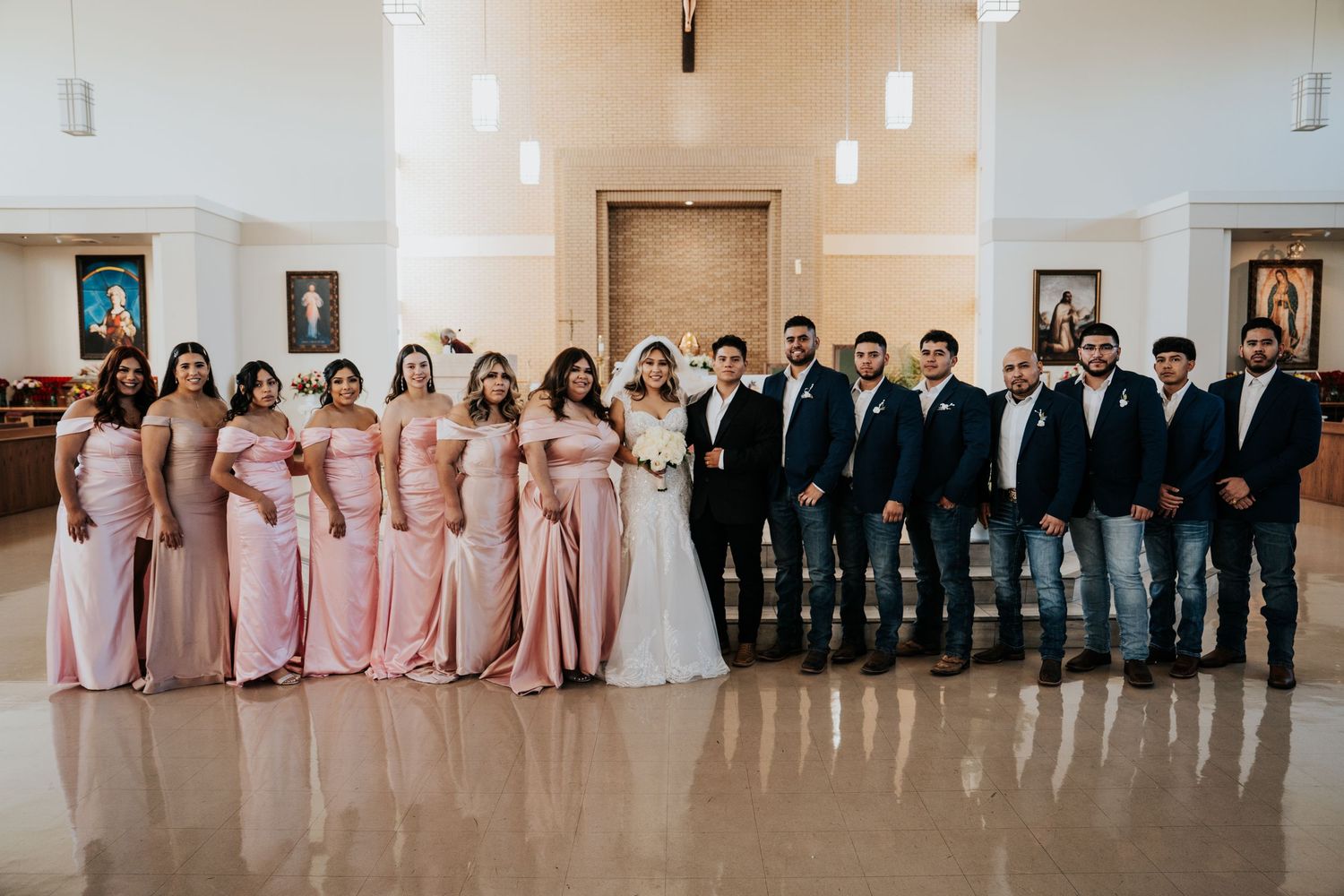 Wedding party members lined up against white wall in alternating pink dresses and dark suits for formal photos.