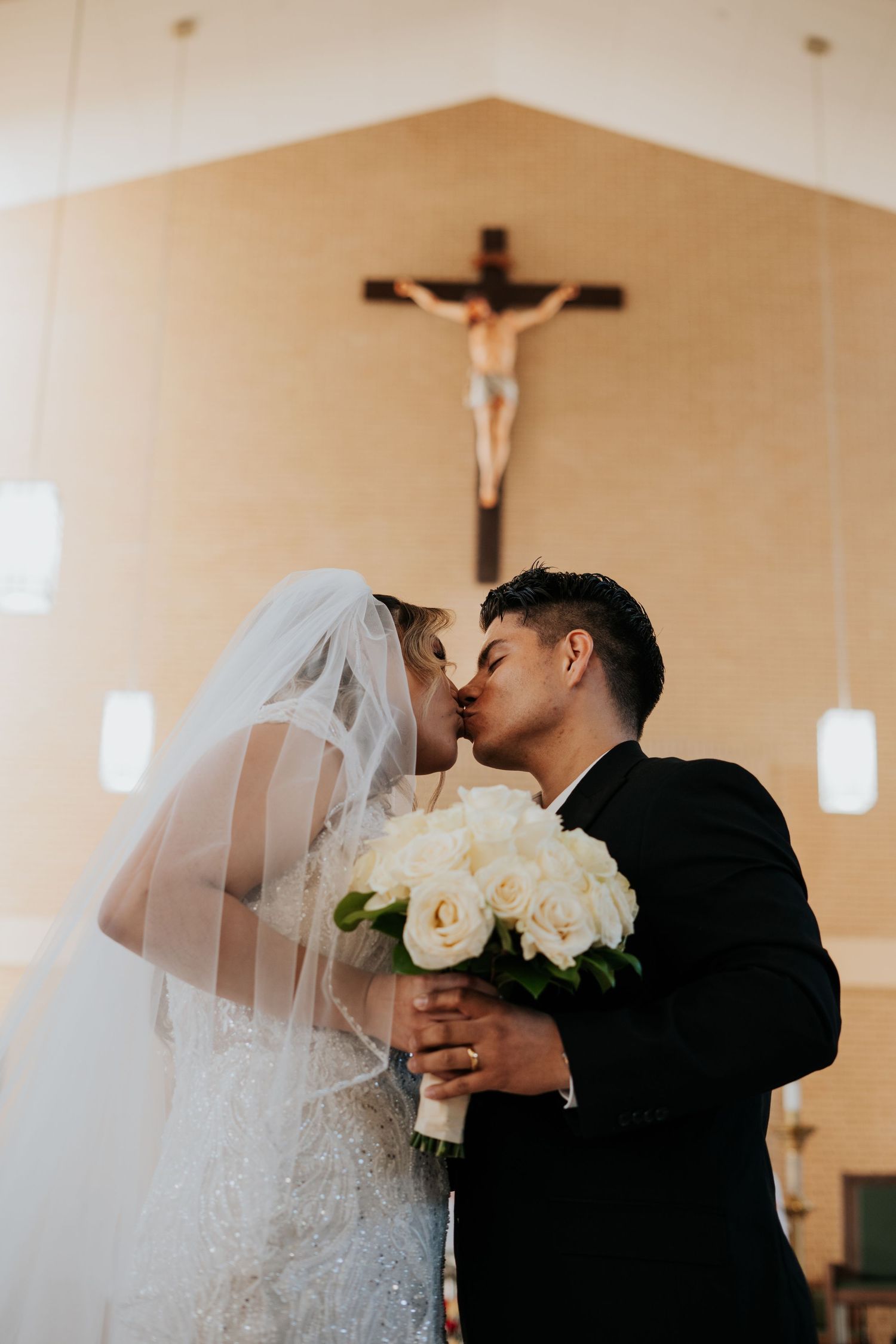 A bride and groom share a romantic kiss in front of a church altar with a crucifix hanging on the wall.