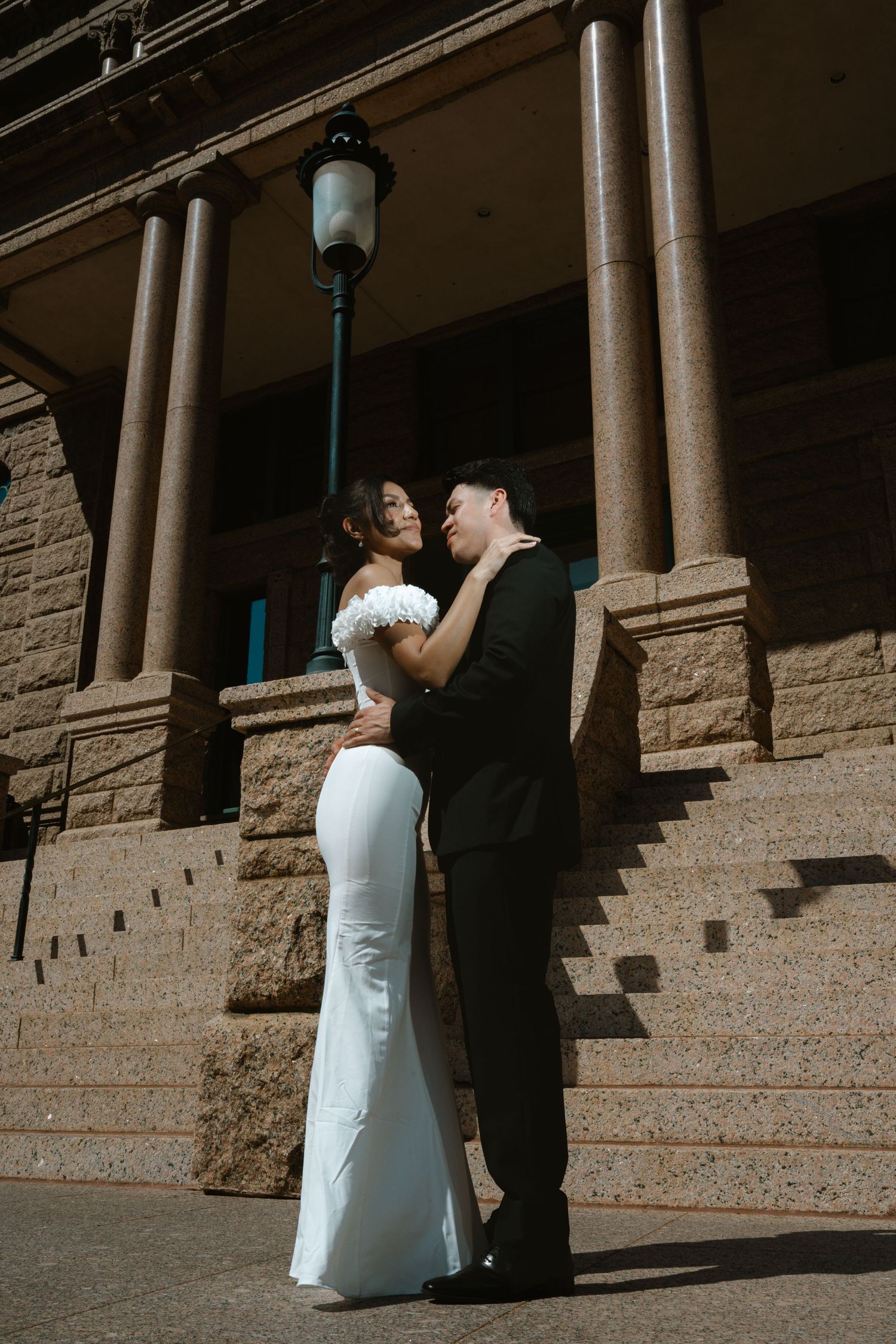 Bride in white gown and groom in black suit embrace near stone columns and lamppost at elegant wedding venue.