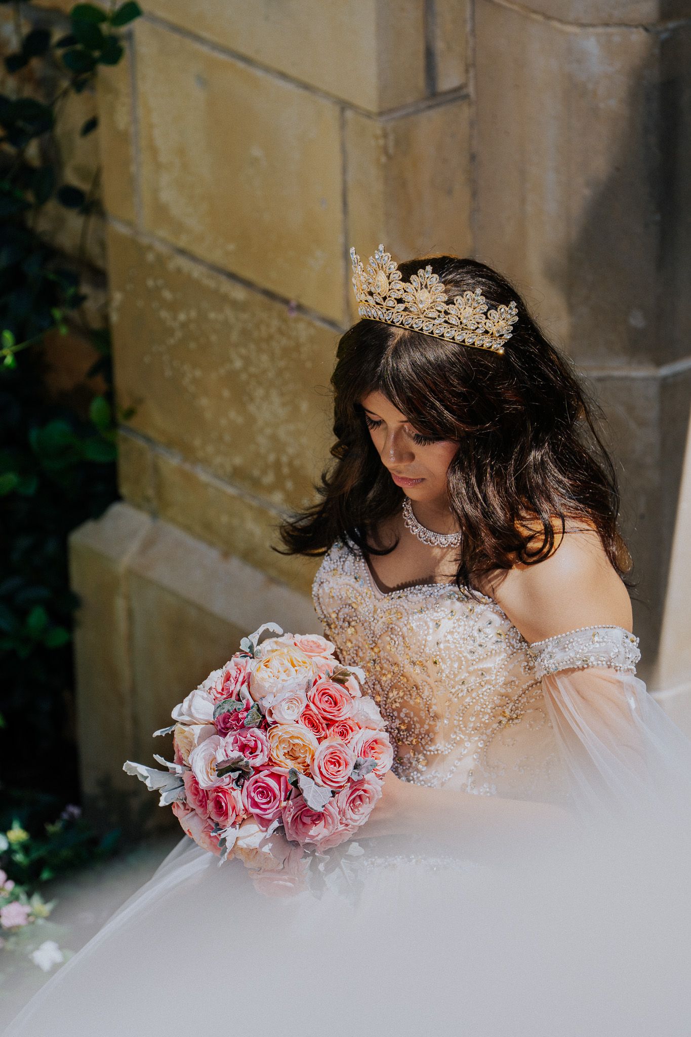 Bride in gold crown and beaded off-shoulder gown holding pink rose bouquet beside stone wall.