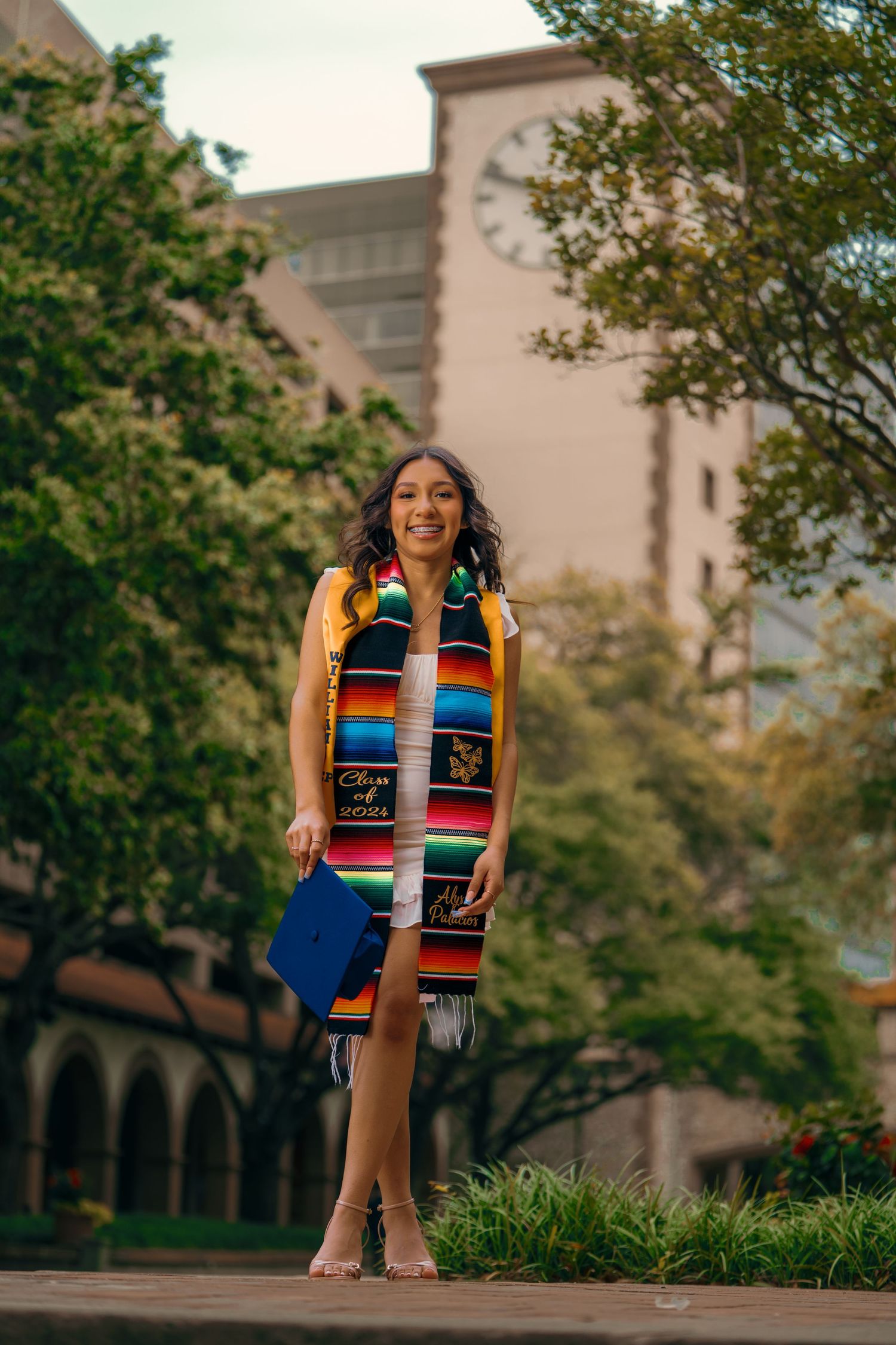 Graduate in blue cap and gown with vibrant Mexican serape stole poses outdoors on campus grounds.