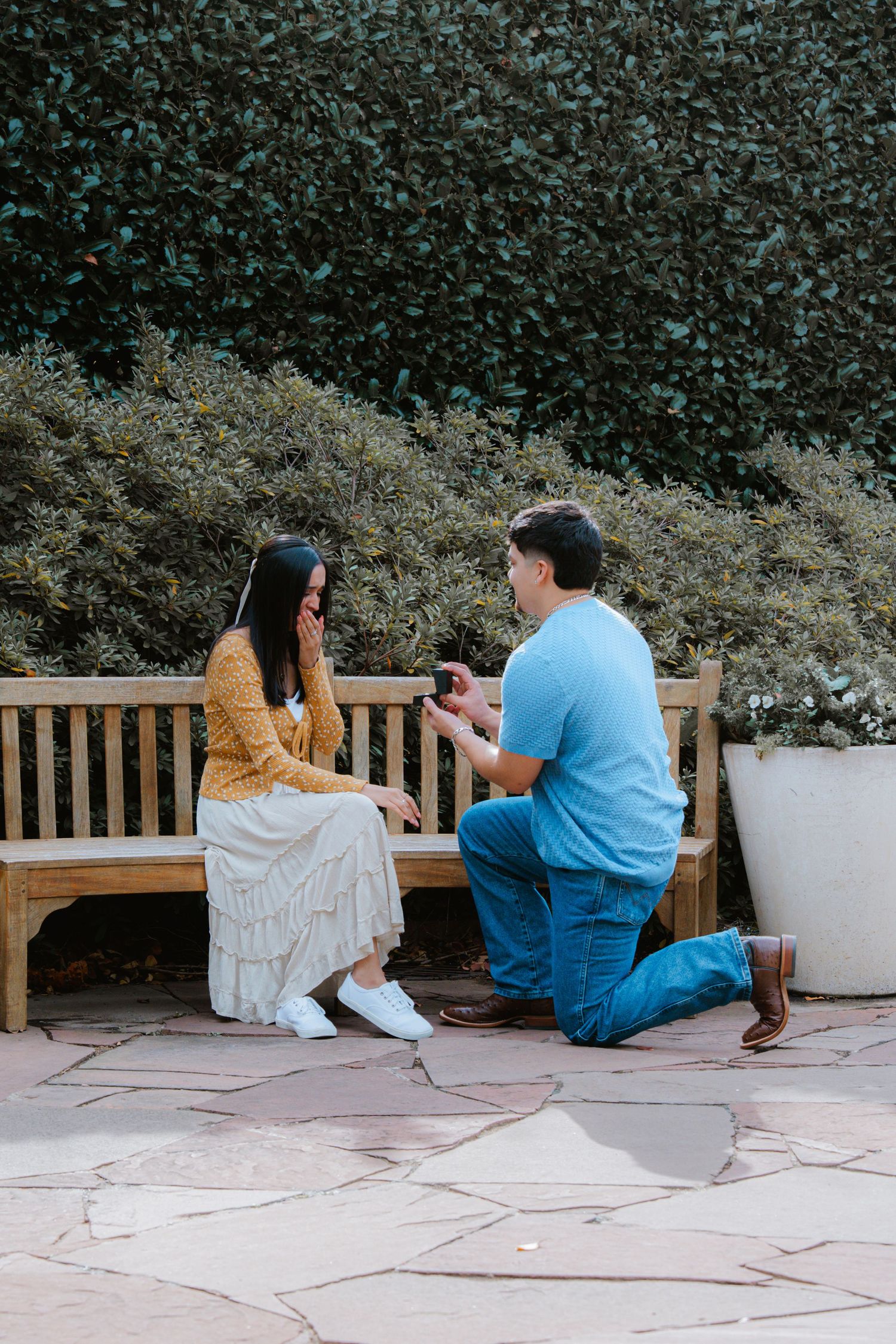 A couple sits together on a wooden bench in front of a hedge, sharing an intimate moment.