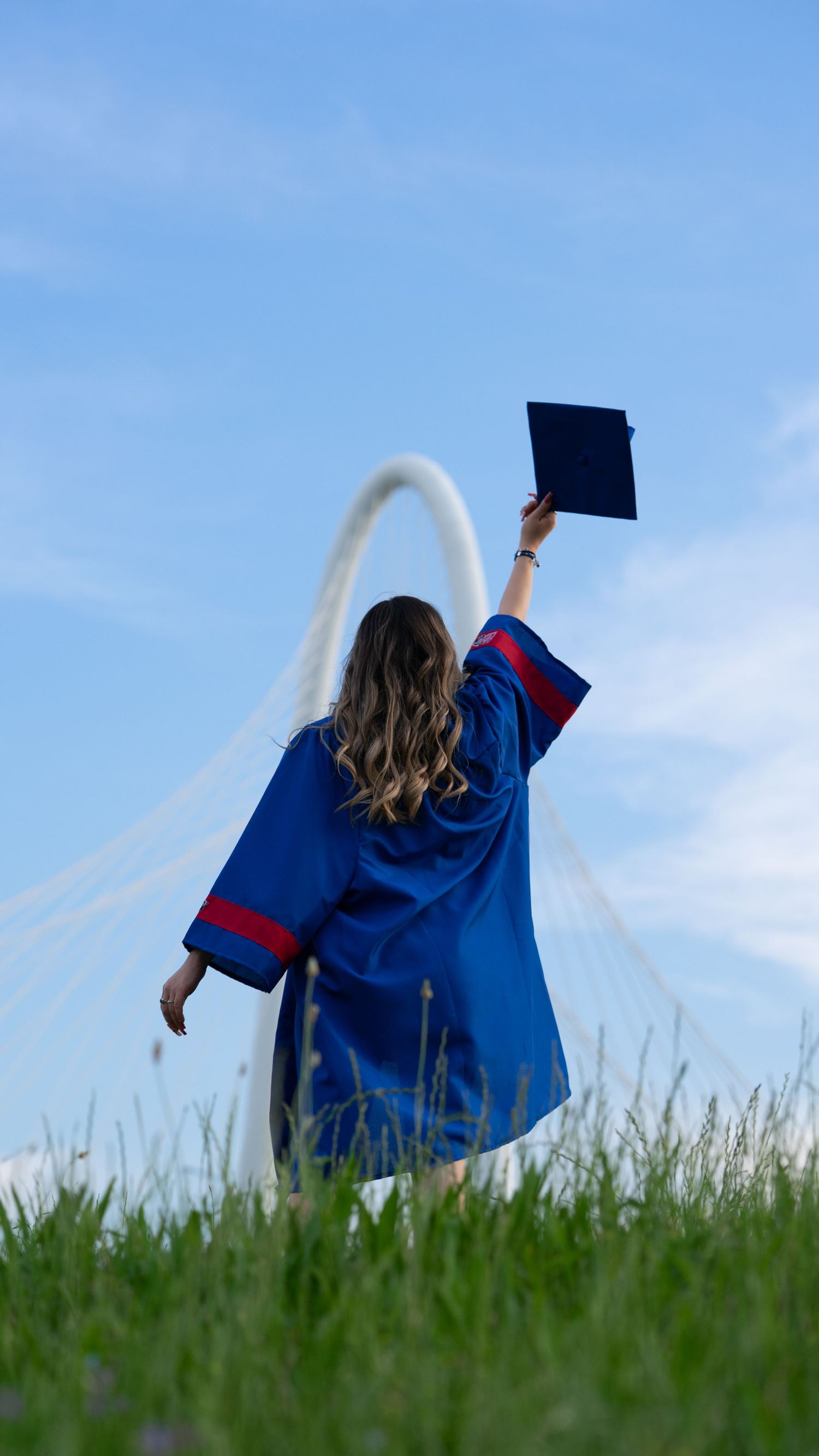 A graduate in blue and red academic regalia celebrates on a grassy hillside under clear skies.