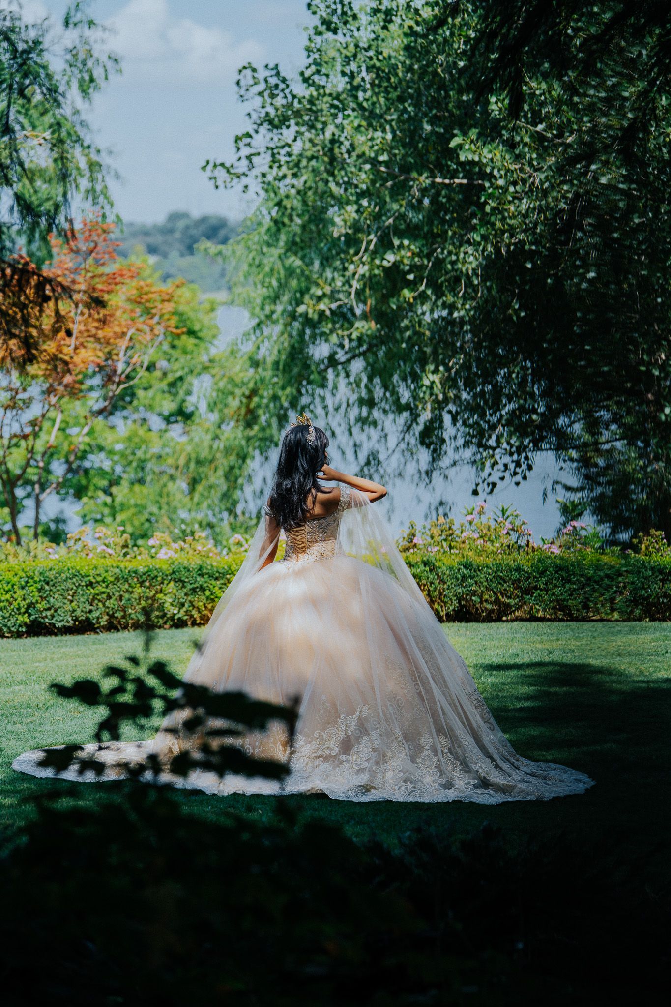 A bride in a flowing ball gown twirls in a lush garden with weeping willow trees and a lake in the background.