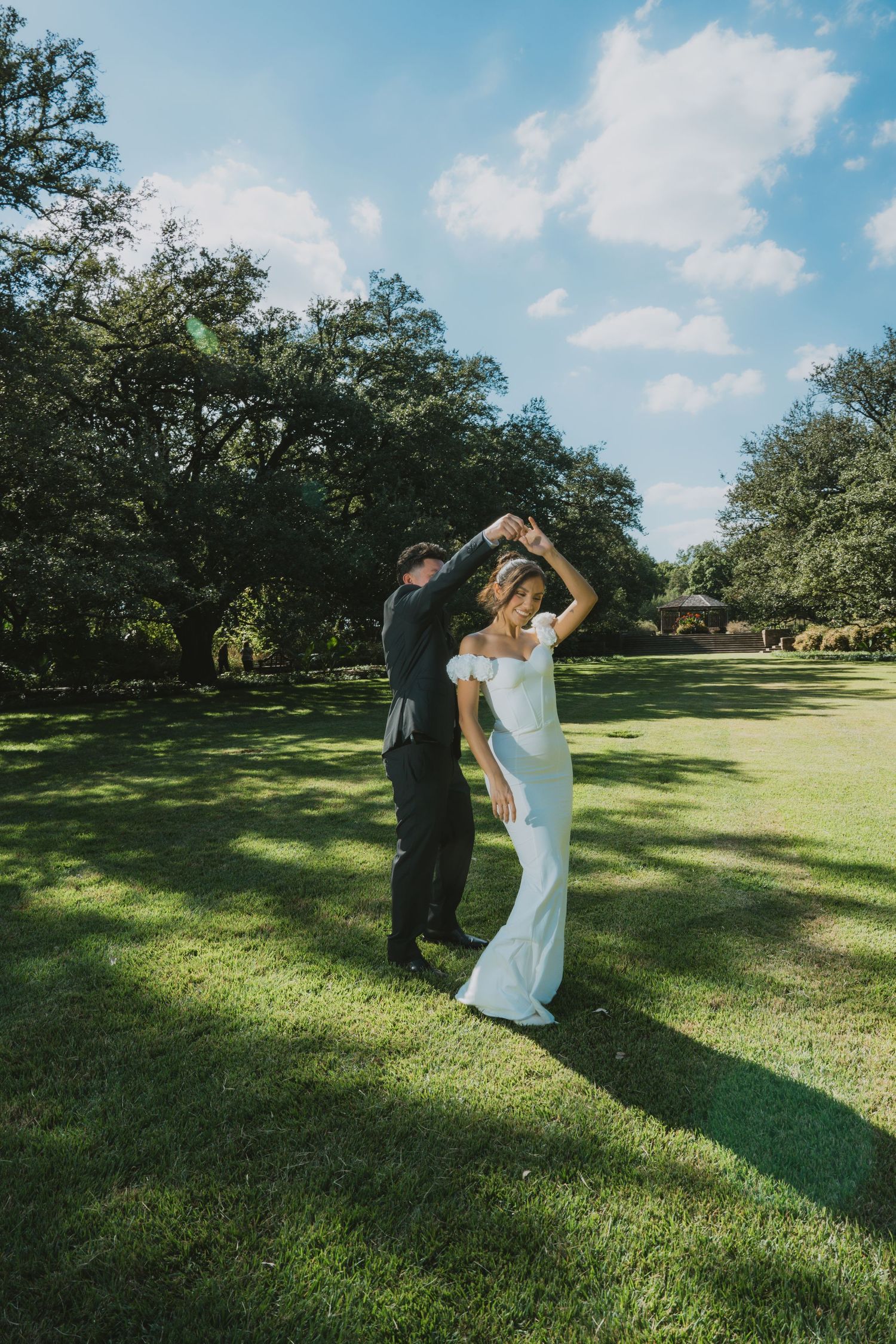Bride in white gown being twirled by groom in black suit on lush green lawn surrounded by trees.