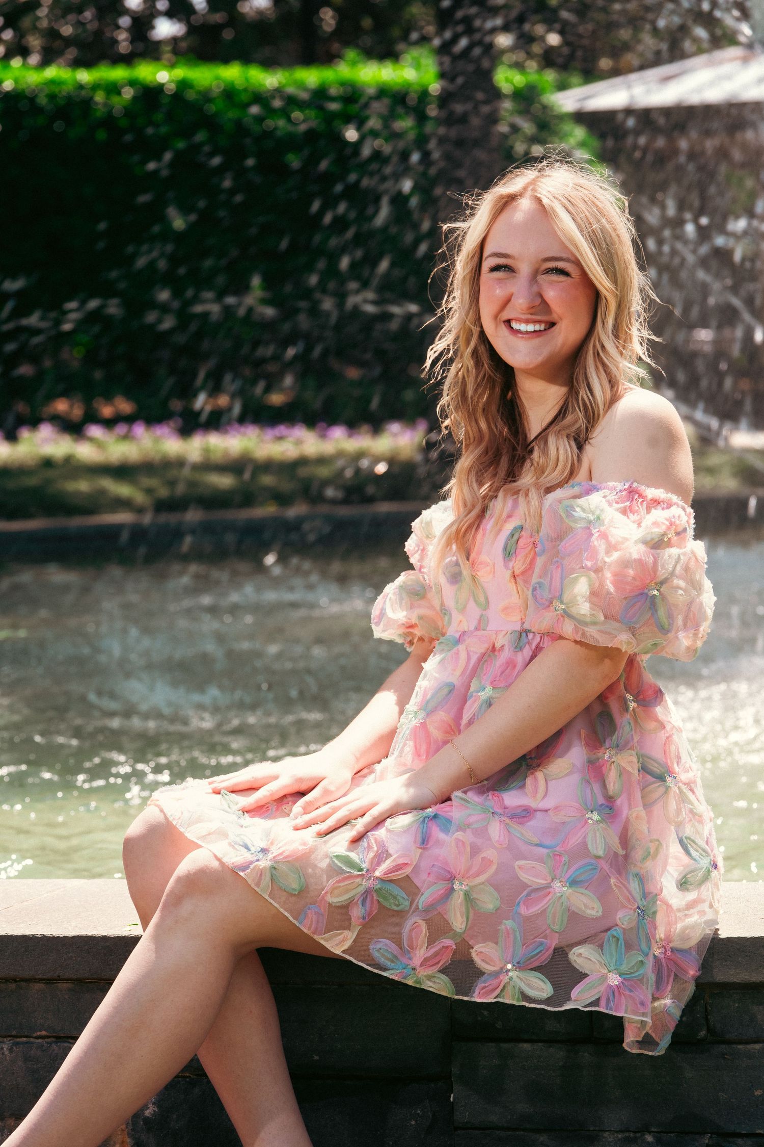 A person in a pink floral dress poses gracefully by a fountain on a sunny summer day.