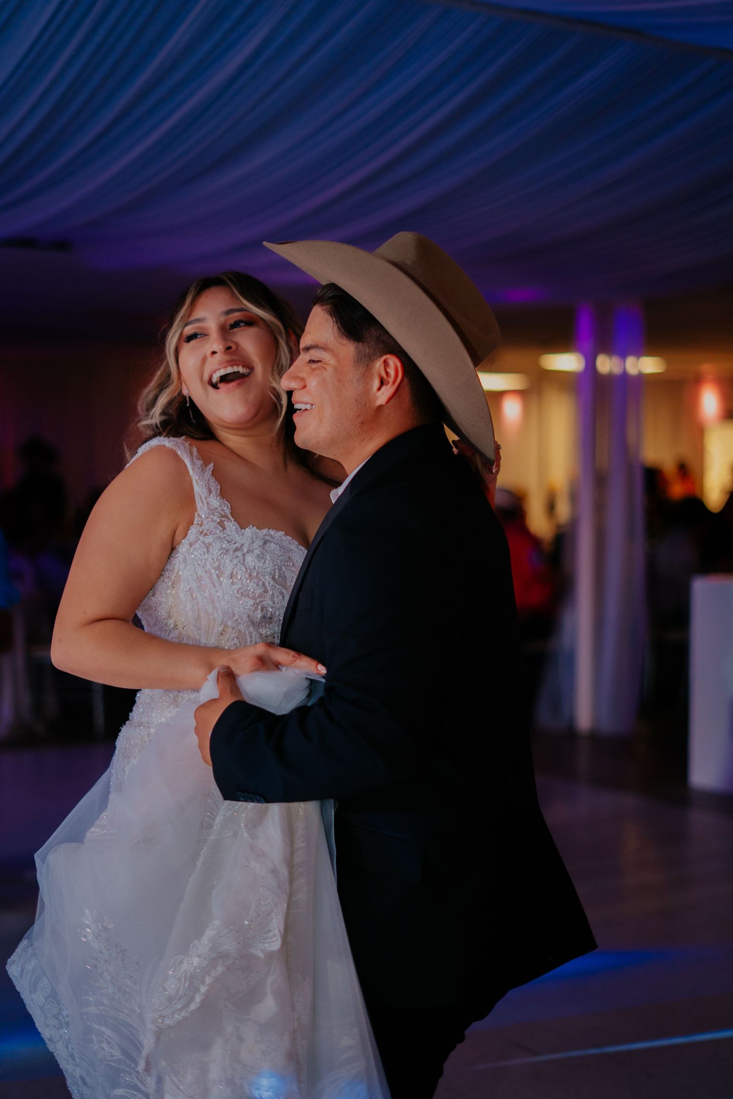 A couple dances and laughs together during their first dance at a wedding reception with blue ambient lighting.