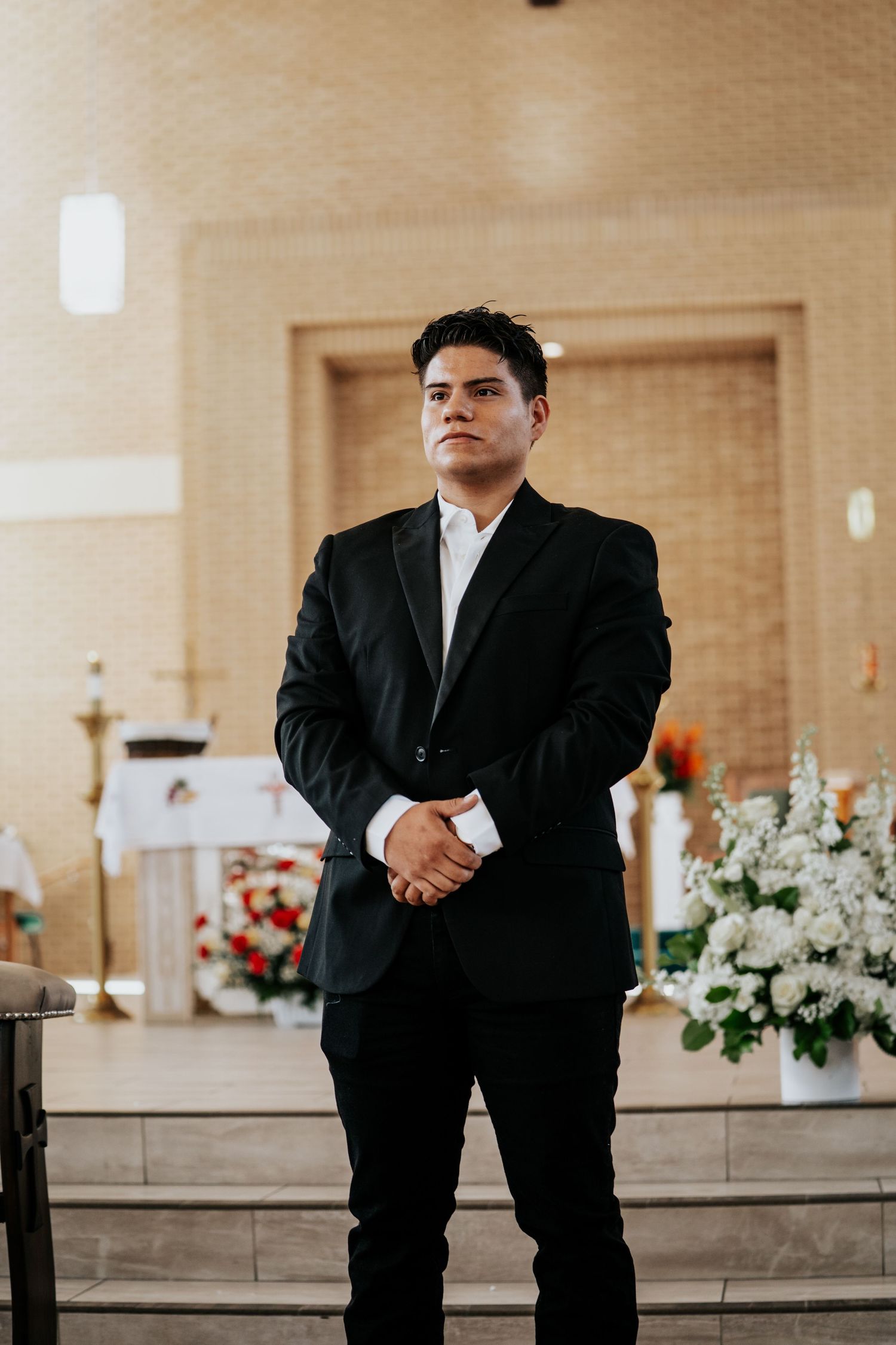 A person in a black suit stands in a church with floral decorations and brick walls in the background.