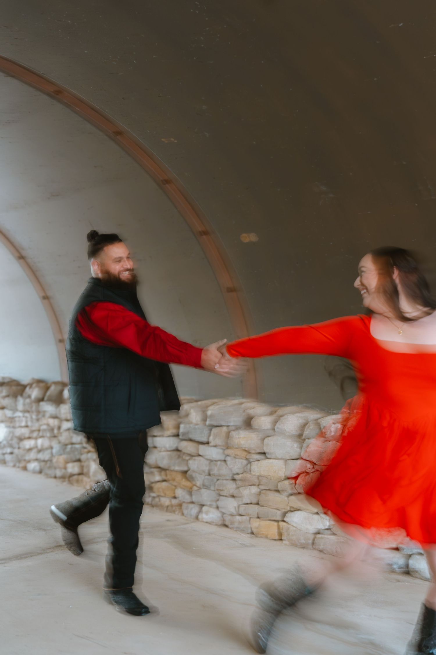 A joyful couple dances in a rustic stone arch venue, woman in red dress and man in black and red outfit.