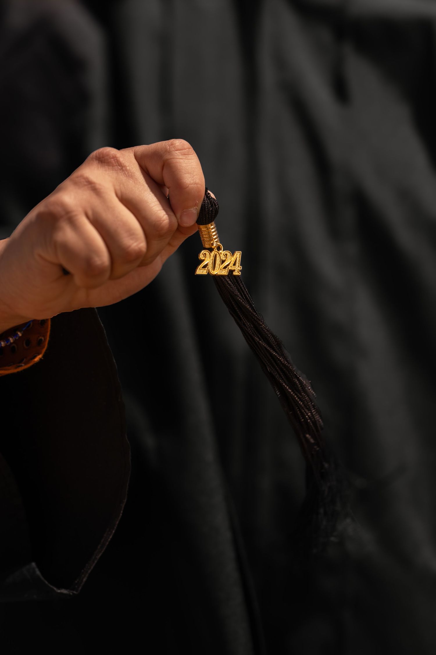 Hand holding 2024 graduation tassel in gold against black graduation gown background.
