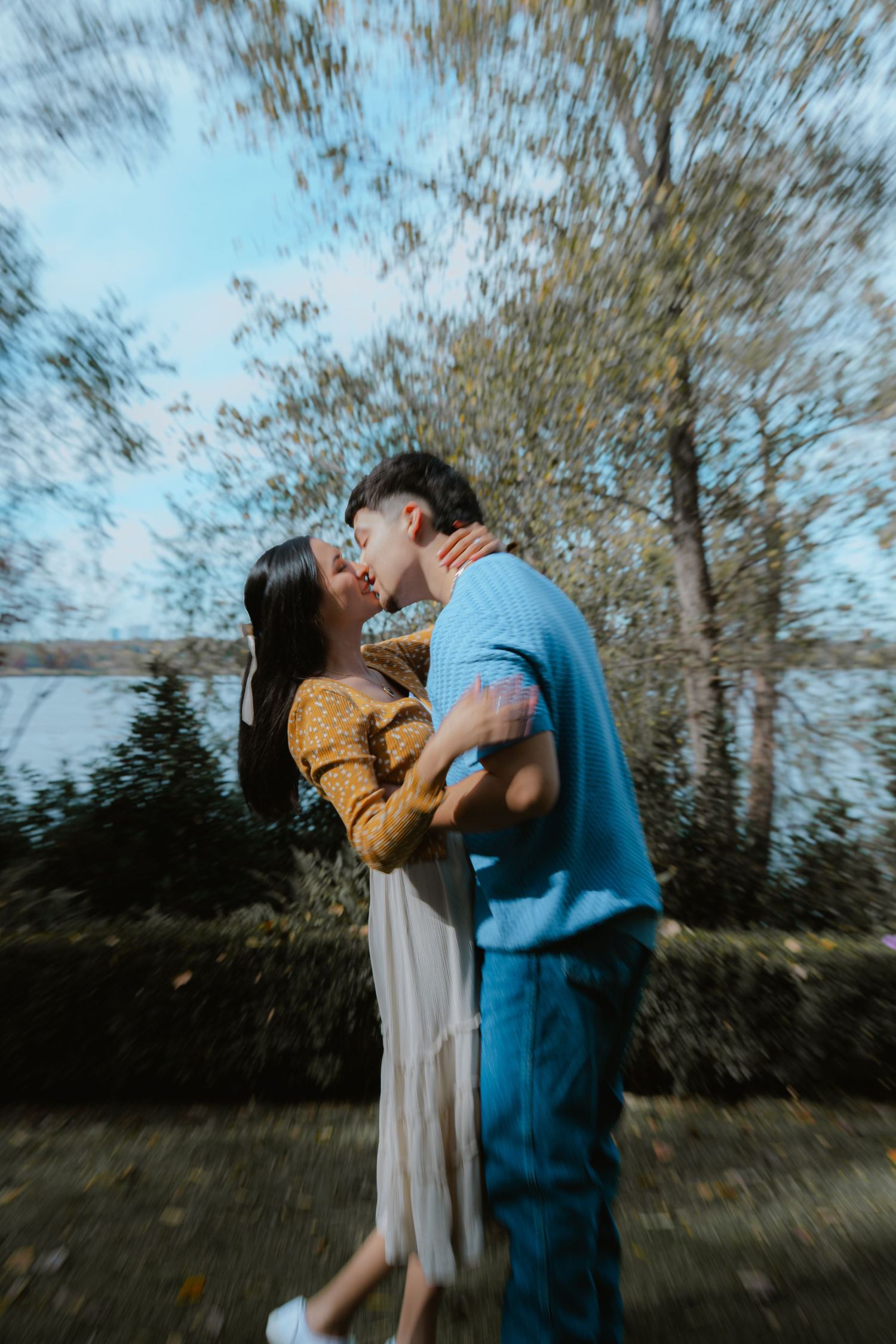 A romantic moment captured between a couple embracing outdoors with trees and water in the background.