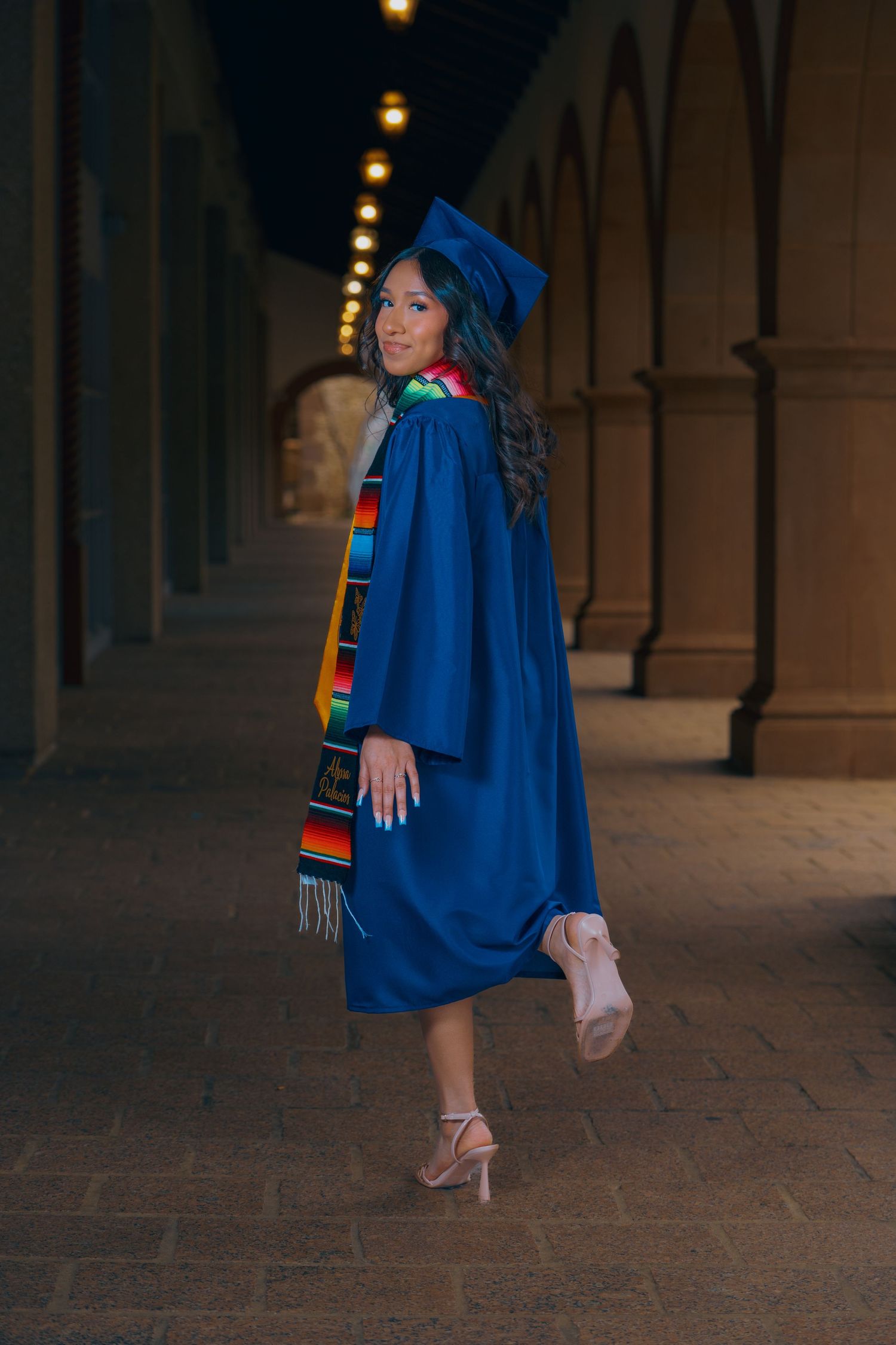 Graduate in blue regalia strikes elegant pose against architectural columns with evening lighting.