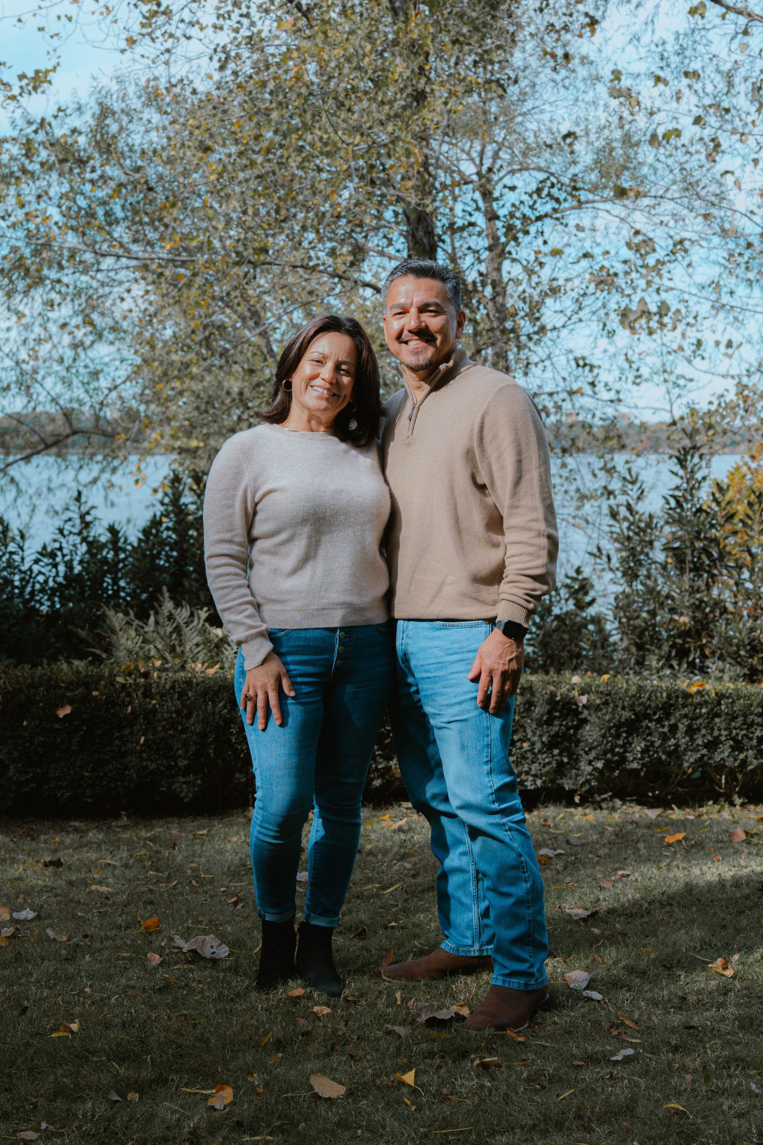 Two people wearing sweaters and jeans pose together outdoors on an autumn day with fallen leaves around them.