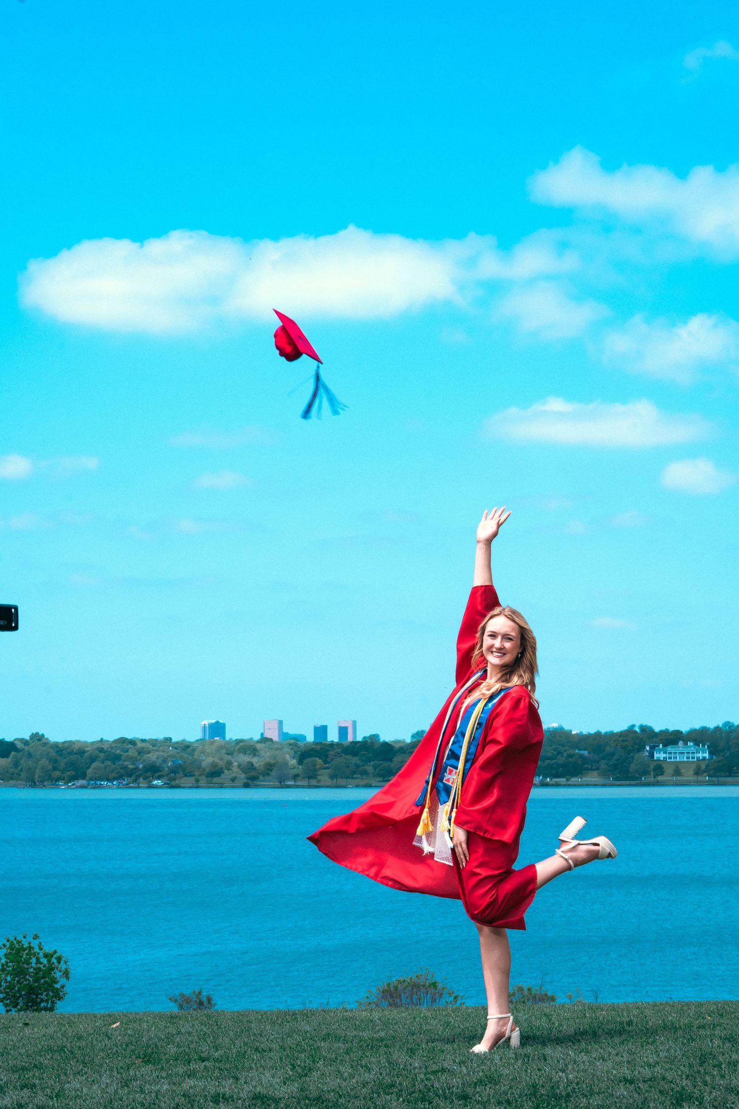 Graduate in red cap and gown celebrates by dancing joyfully on grassy hill overlooking blue lake and city skyline.