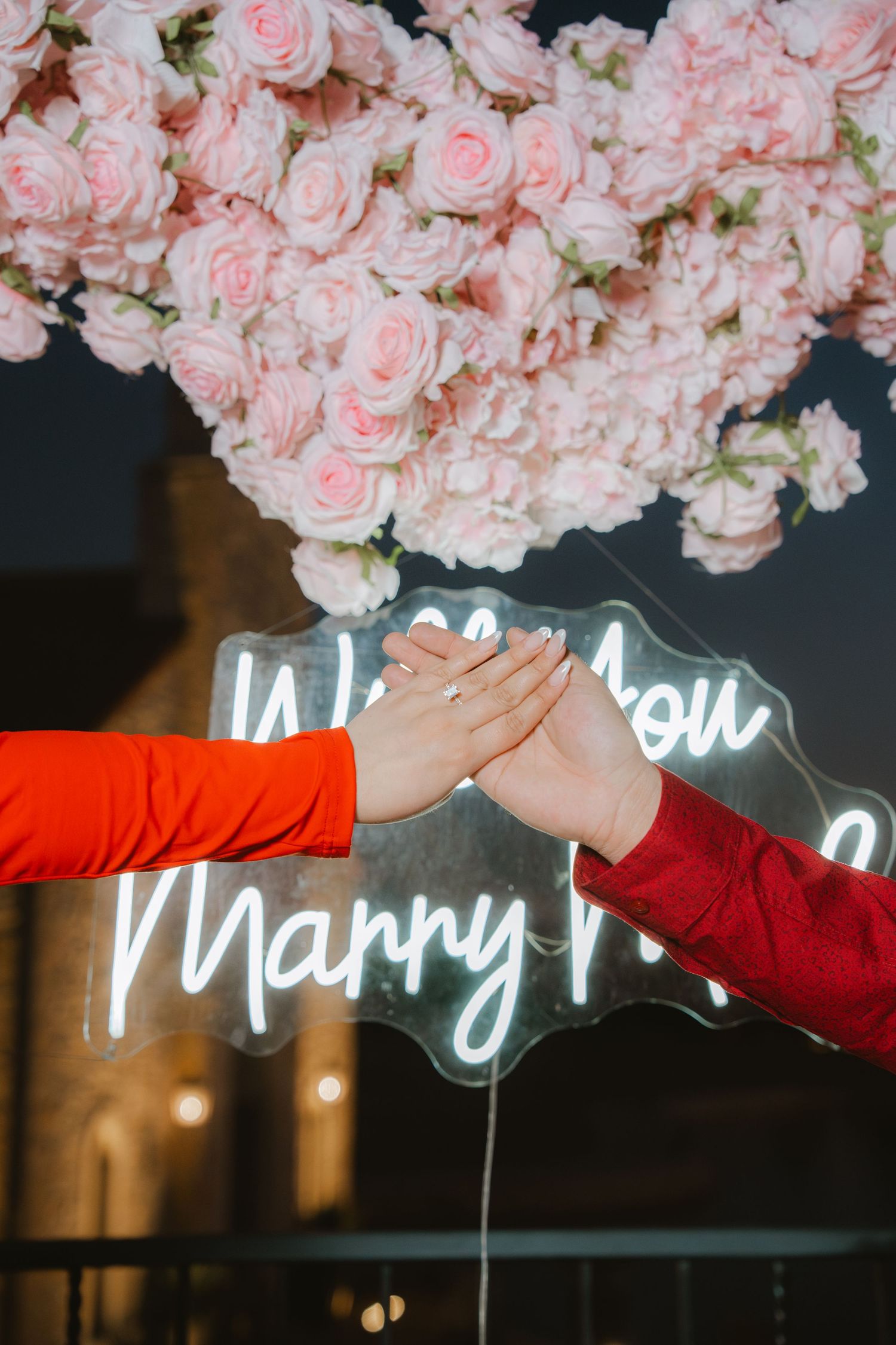 Close-up of hands touching over a neon Will You Marry Me sign surrounded by pink roses at a proposal.