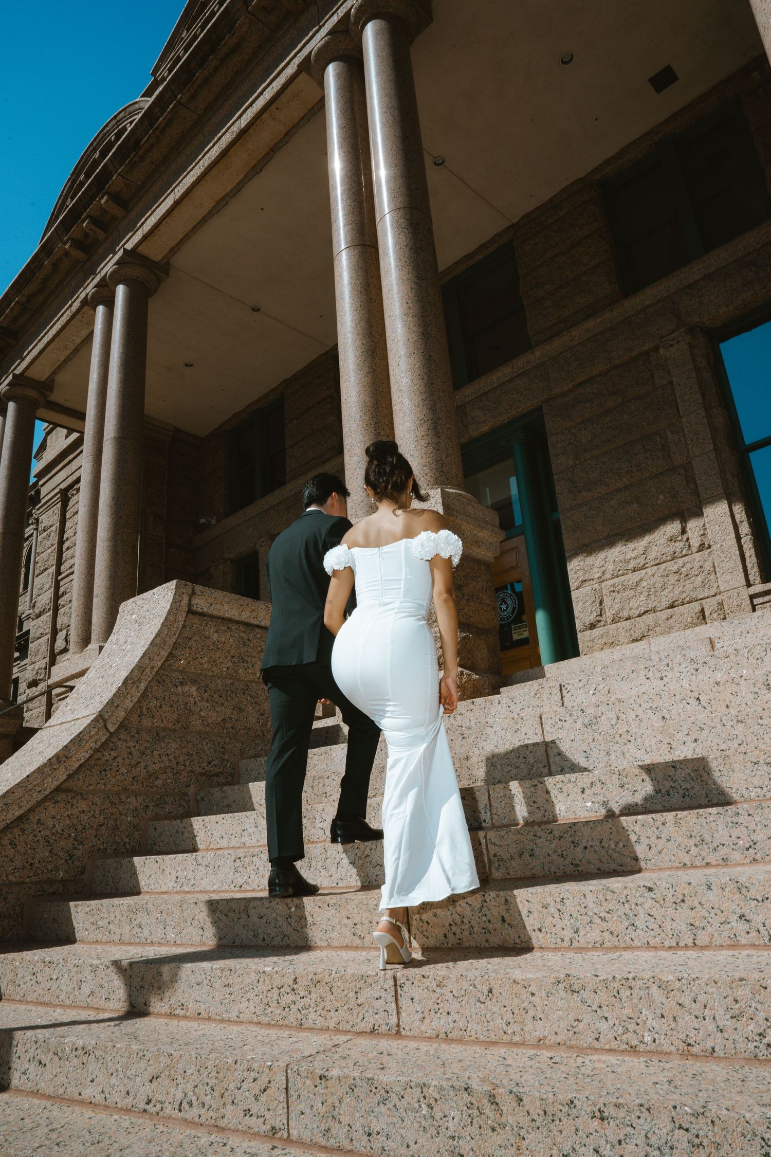 Bride in white off-shoulder gown and groom in dark suit walking up stone steps of elegant columned building.