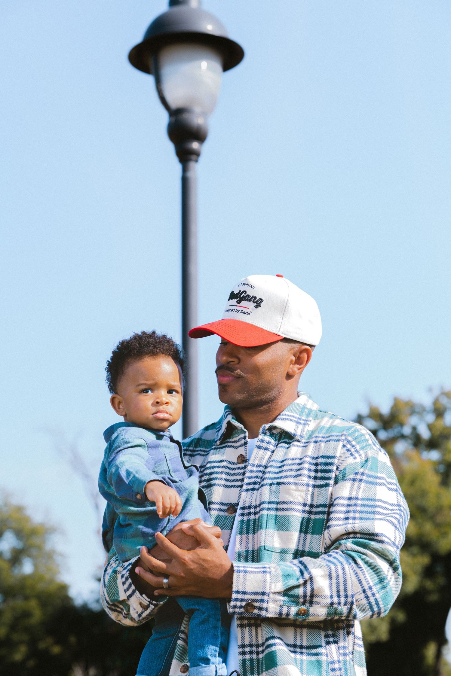 A man in a plaid shirt and cap holds a baby outdoors near a lamp post under a clear blue sky.