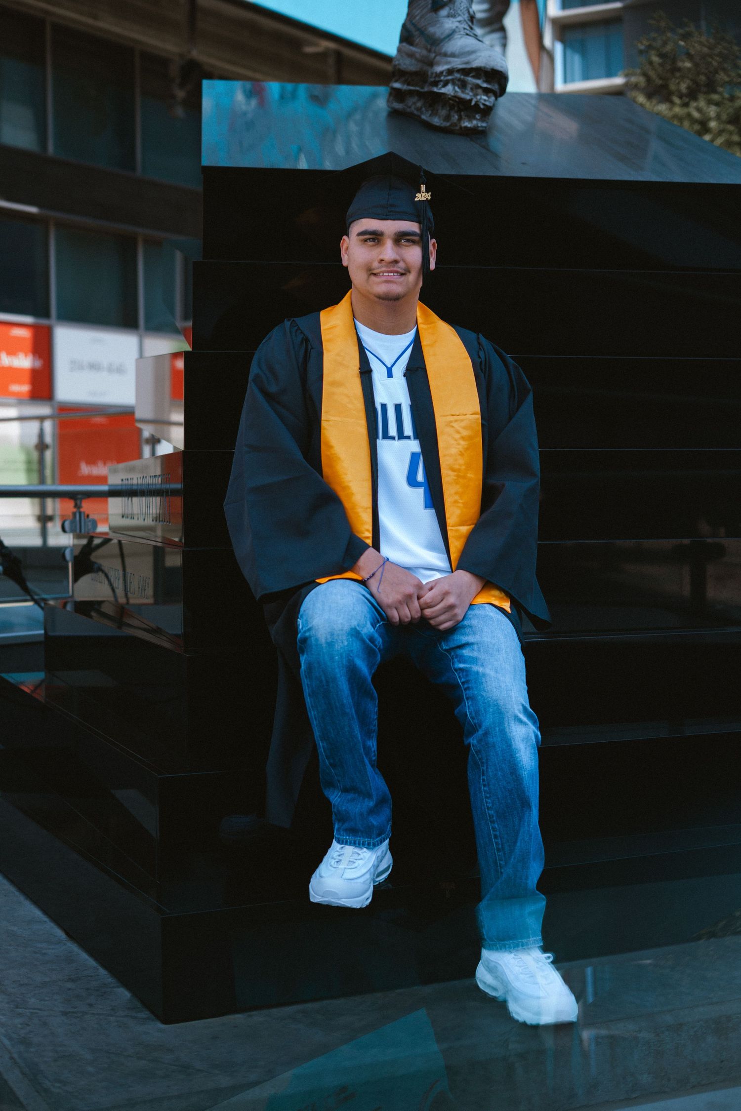 Young man in graduation cap, gown, and gold stole sits smiling outdoors in jeans and white sneakers.