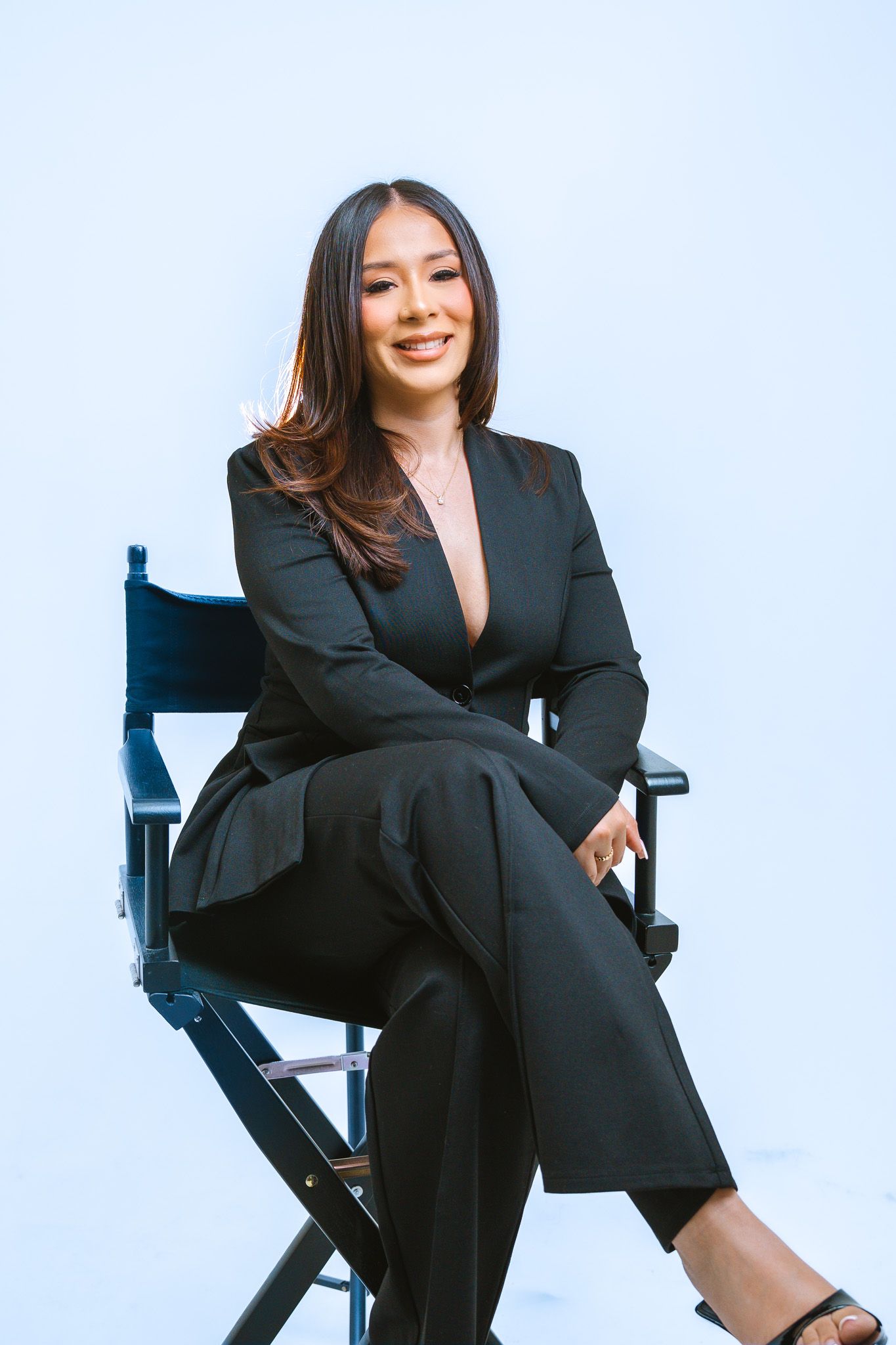 Professional businesswoman in black suit sits confidently in director's chair against light background.