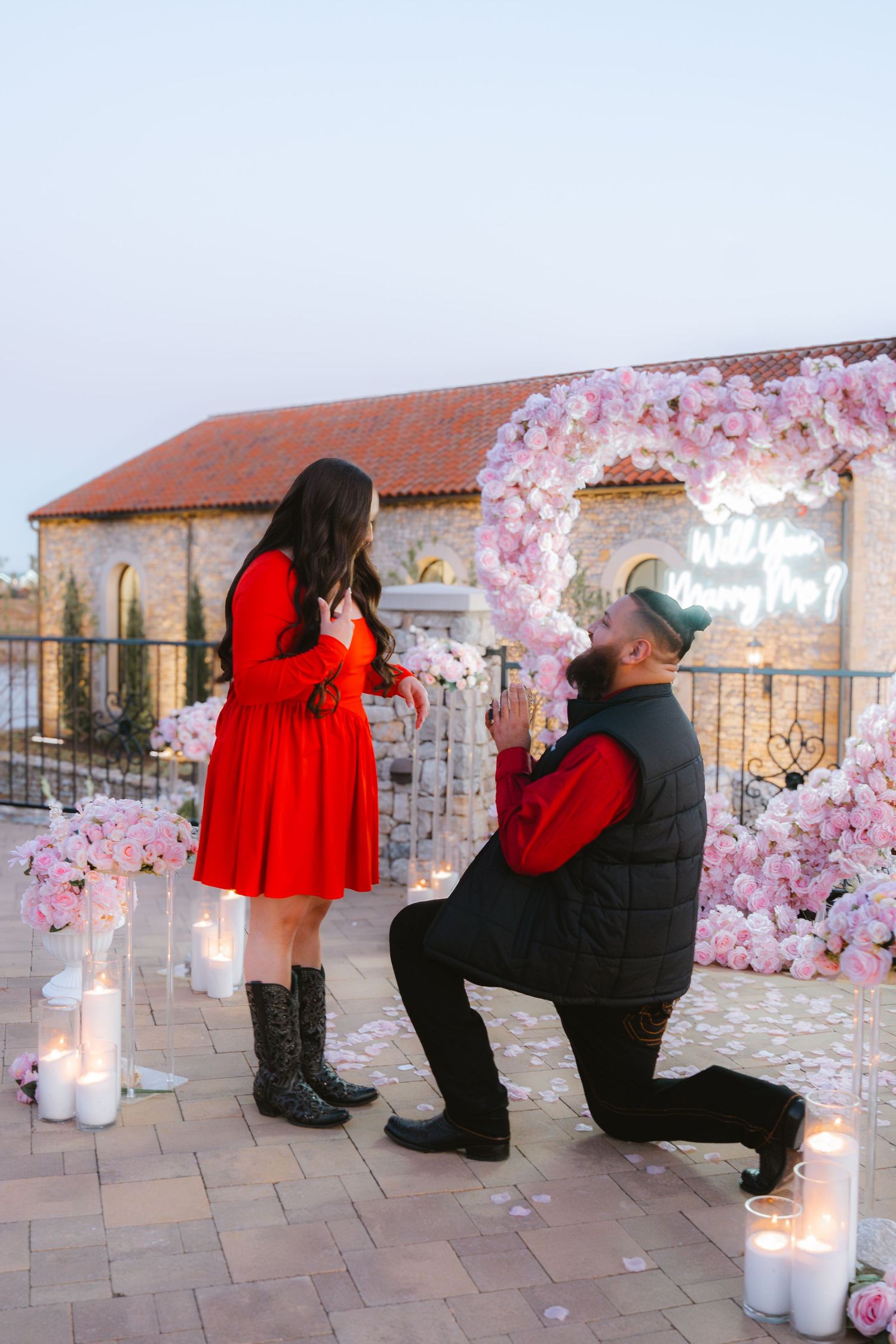 Man proposes to woman in red dress on rooftop surrounded by pink flowers, candles, and a floral heart arch.