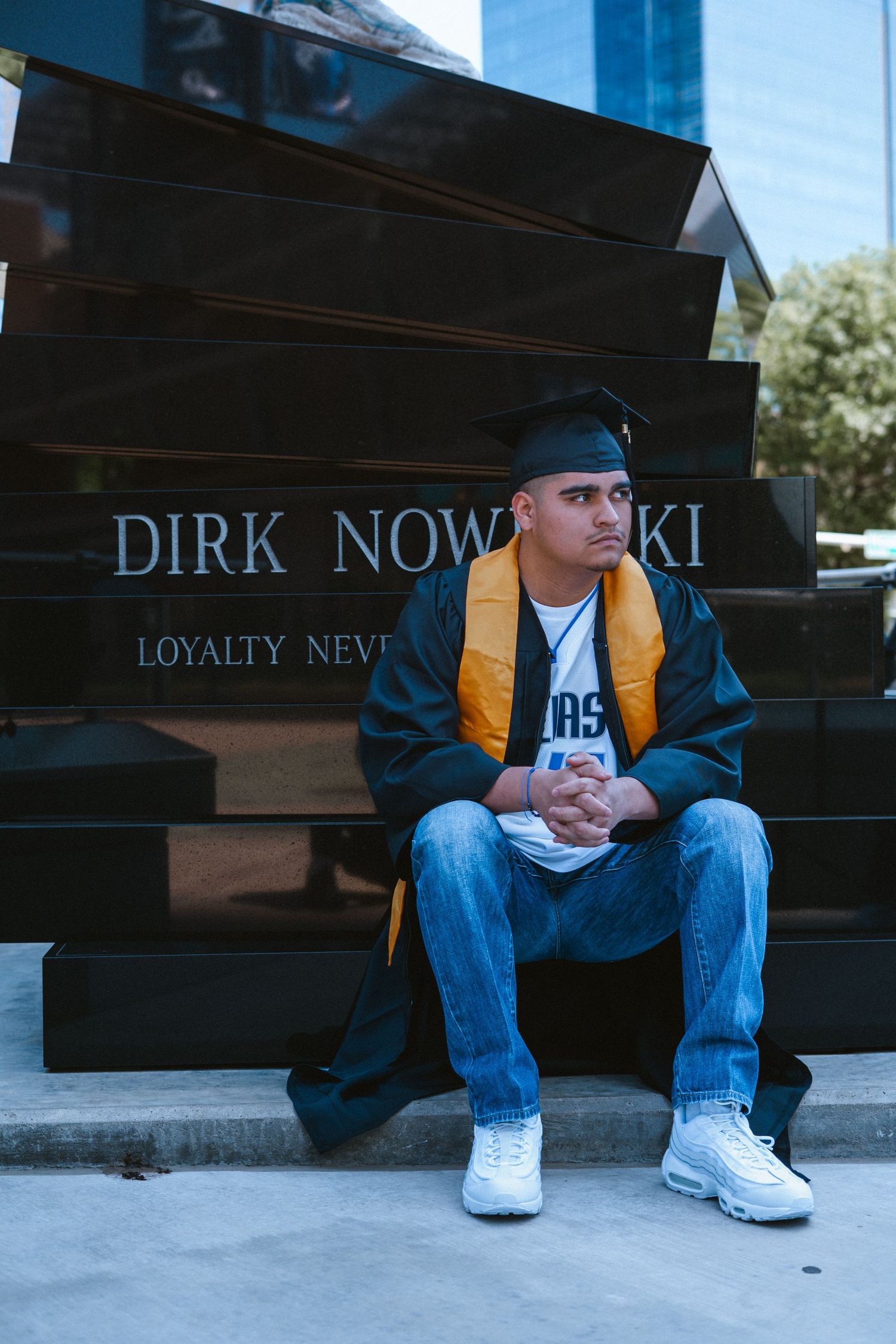 Graduate poses in cap and gown beside Dirk Nowitzki monument with Loyalty Never inscription in Dallas.