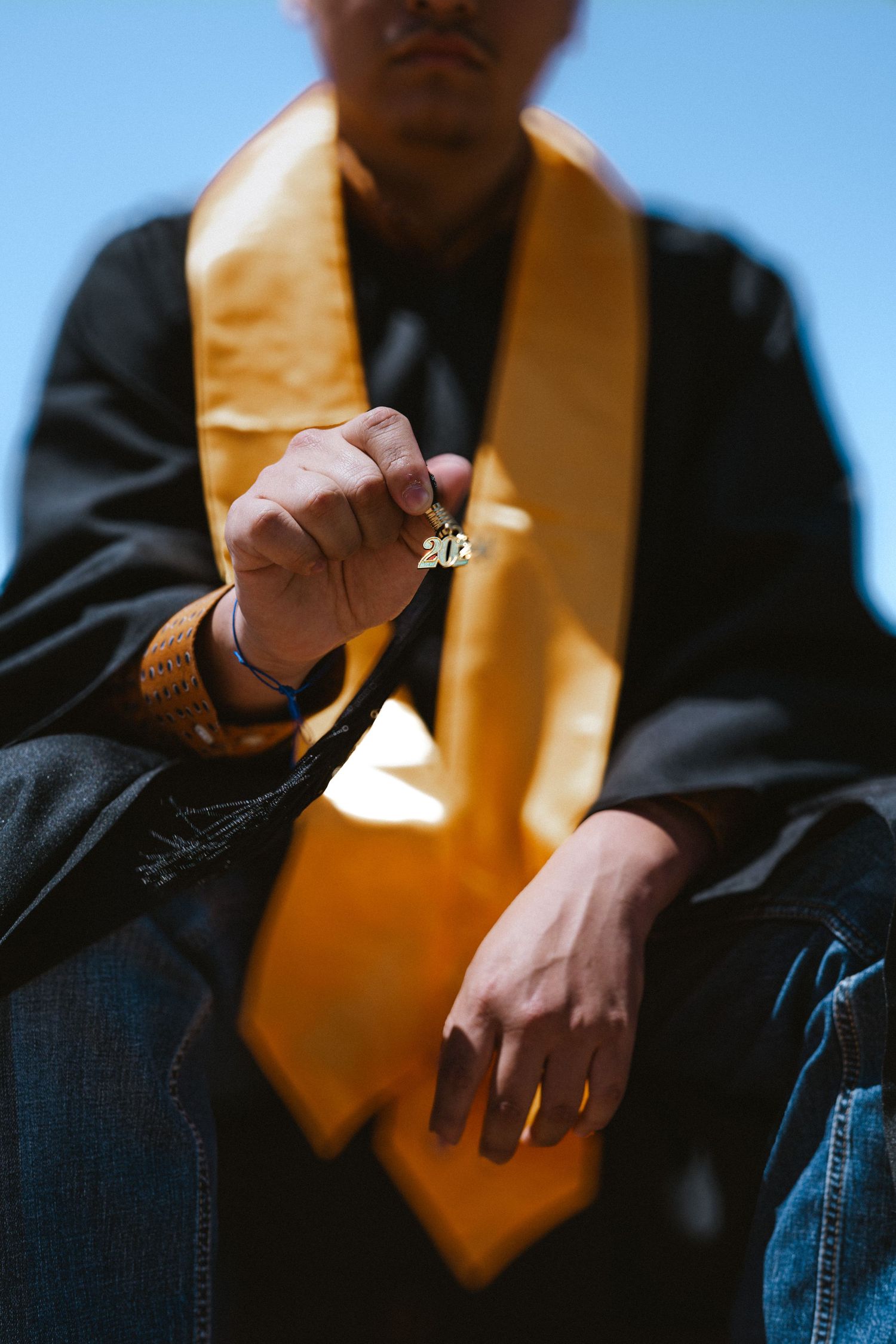 Graduate in black cap and gold stole holds class ring toward camera against blue sky.