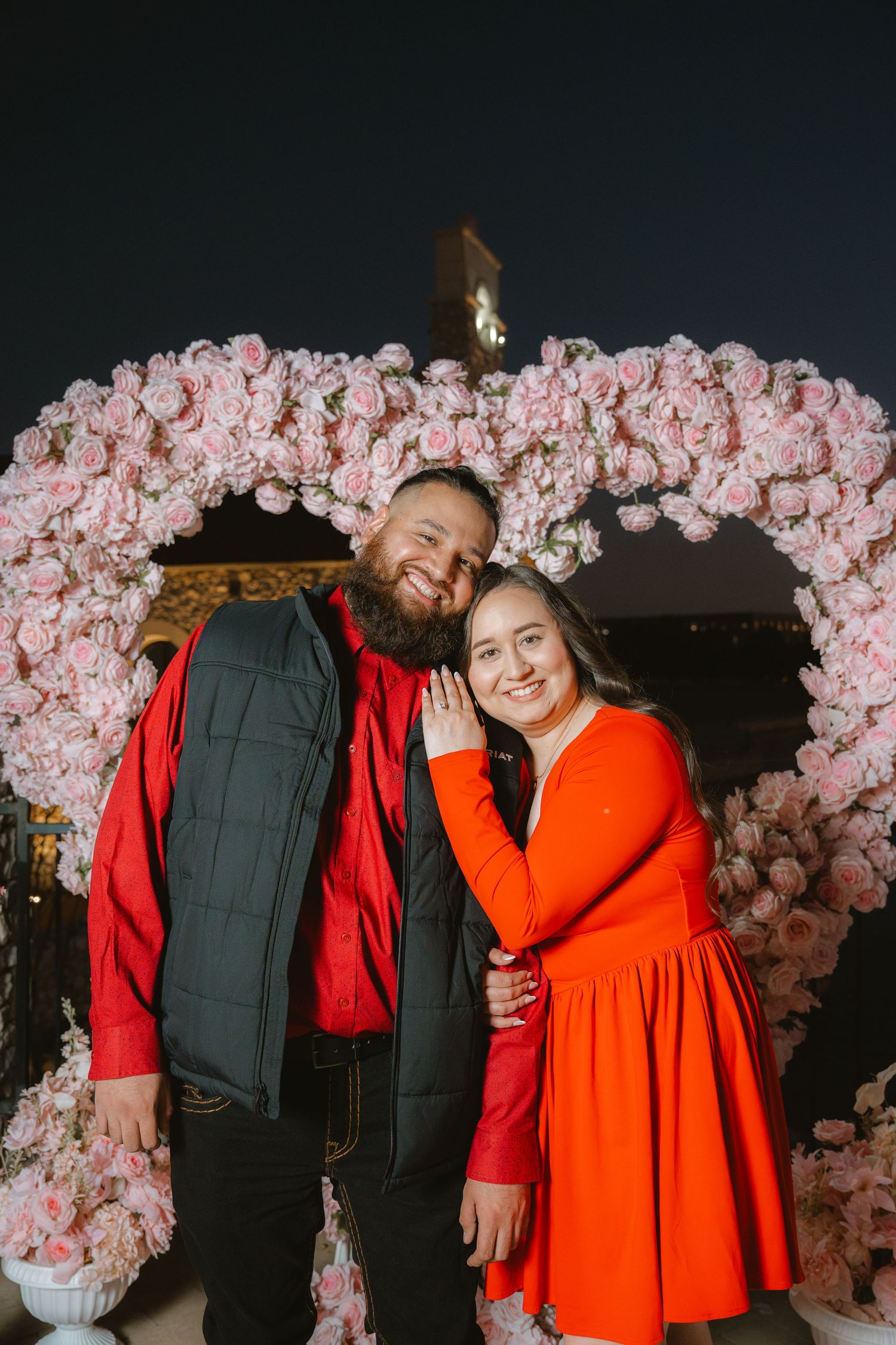 Smiling engaged couple posing together in front of a large pink floral heart arch at night.