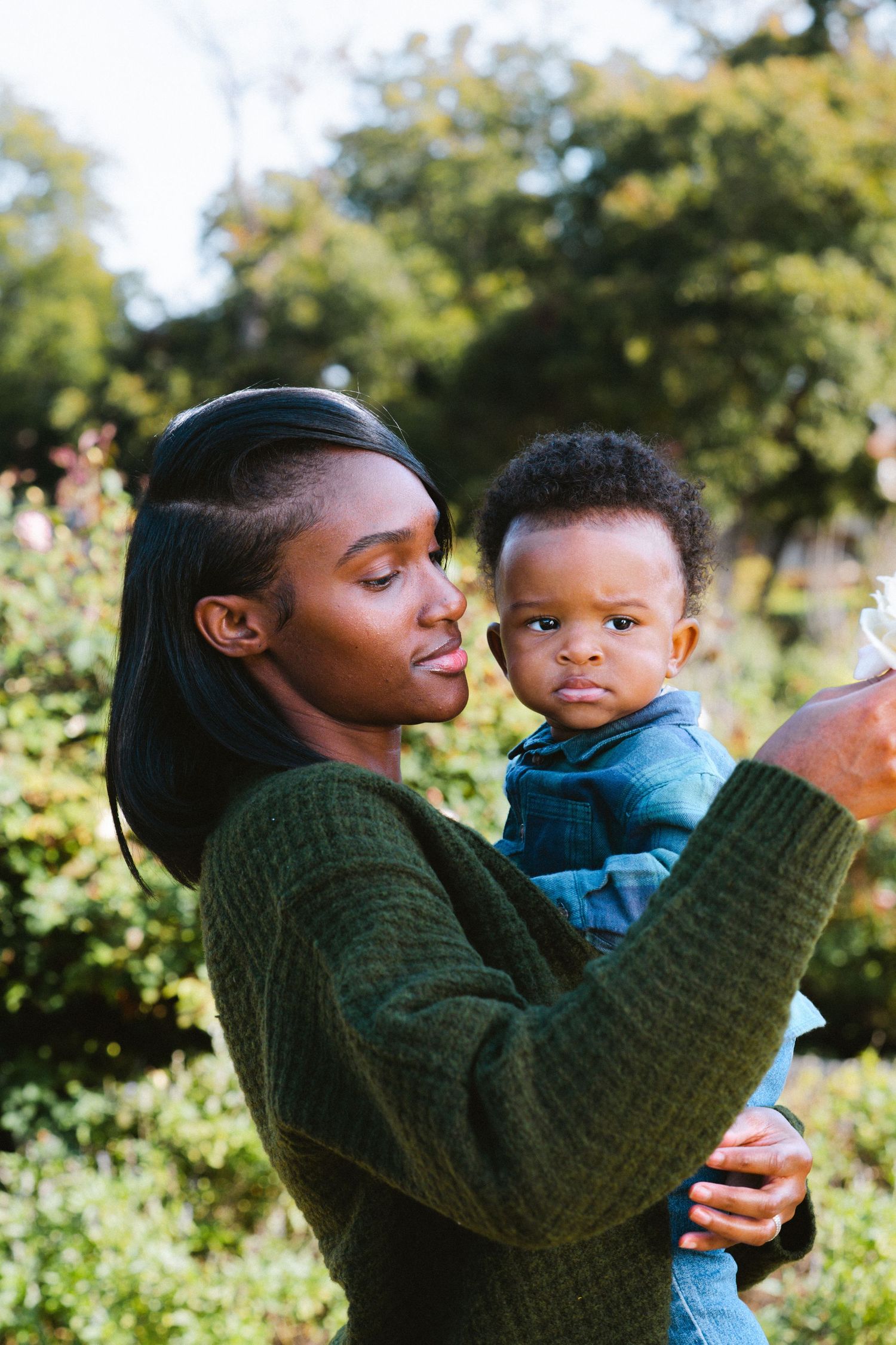Young Black mother in green sweater tenderly holding her baby boy outdoors in a lush garden setting.