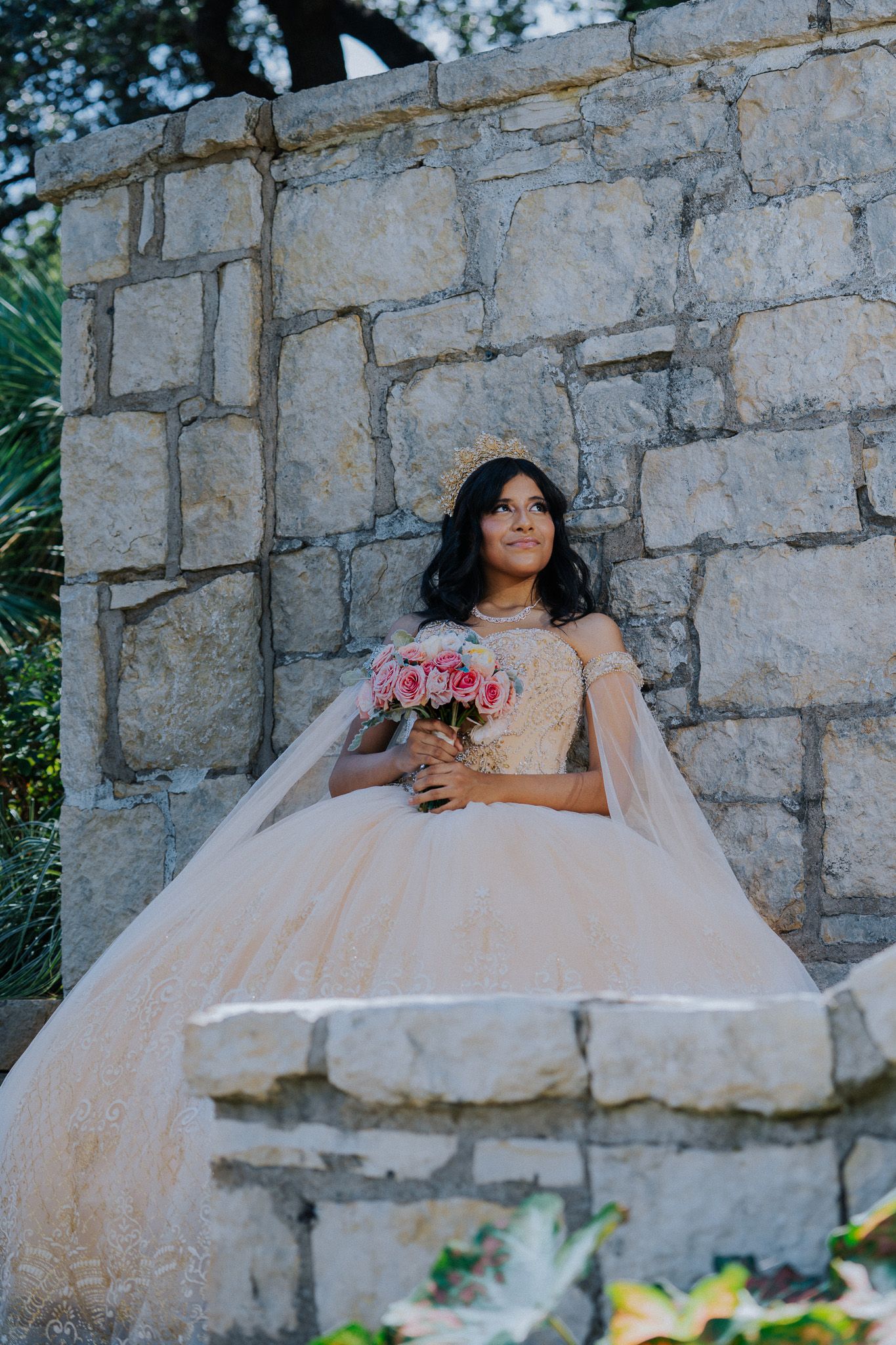Young woman in blush ball gown holding pink bouquet poses by stone wall during quinceañera celebration.