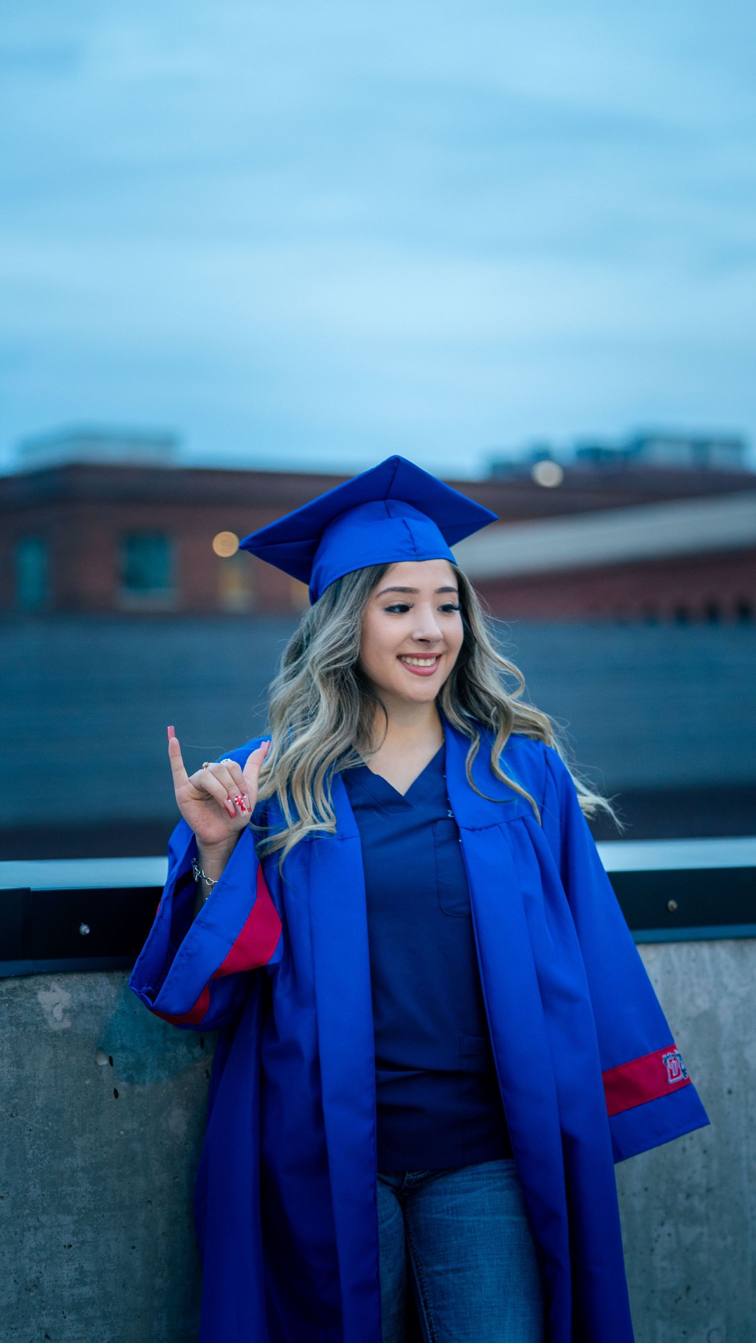 A graduate in a royal blue cap and gown poses outdoors against a blue sky background.