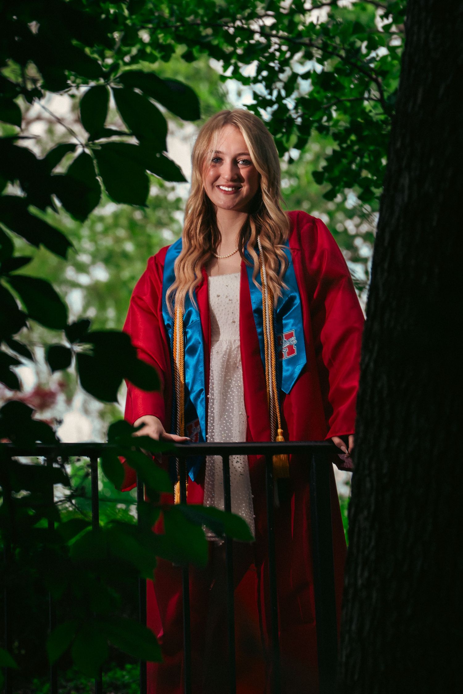 Graduate in red academic regalia poses outdoors among green foliage for graduation photos.