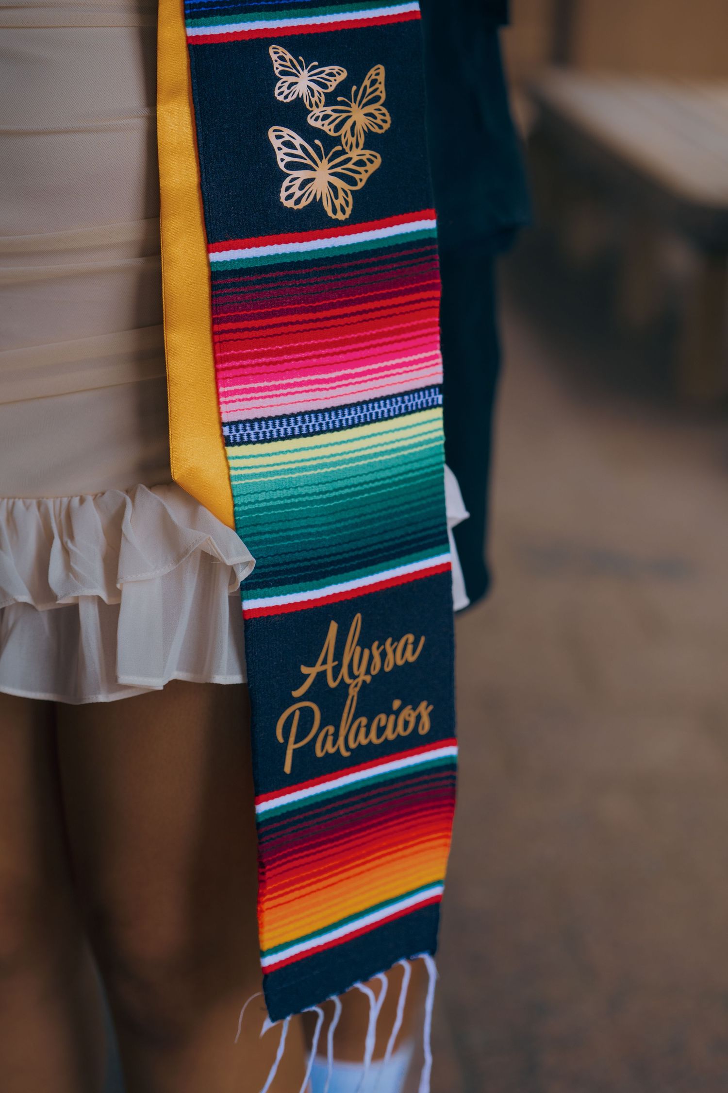 Colorful Mexican serape graduation stoles with gold embroidered text and butterfly details displayed against white fabric.