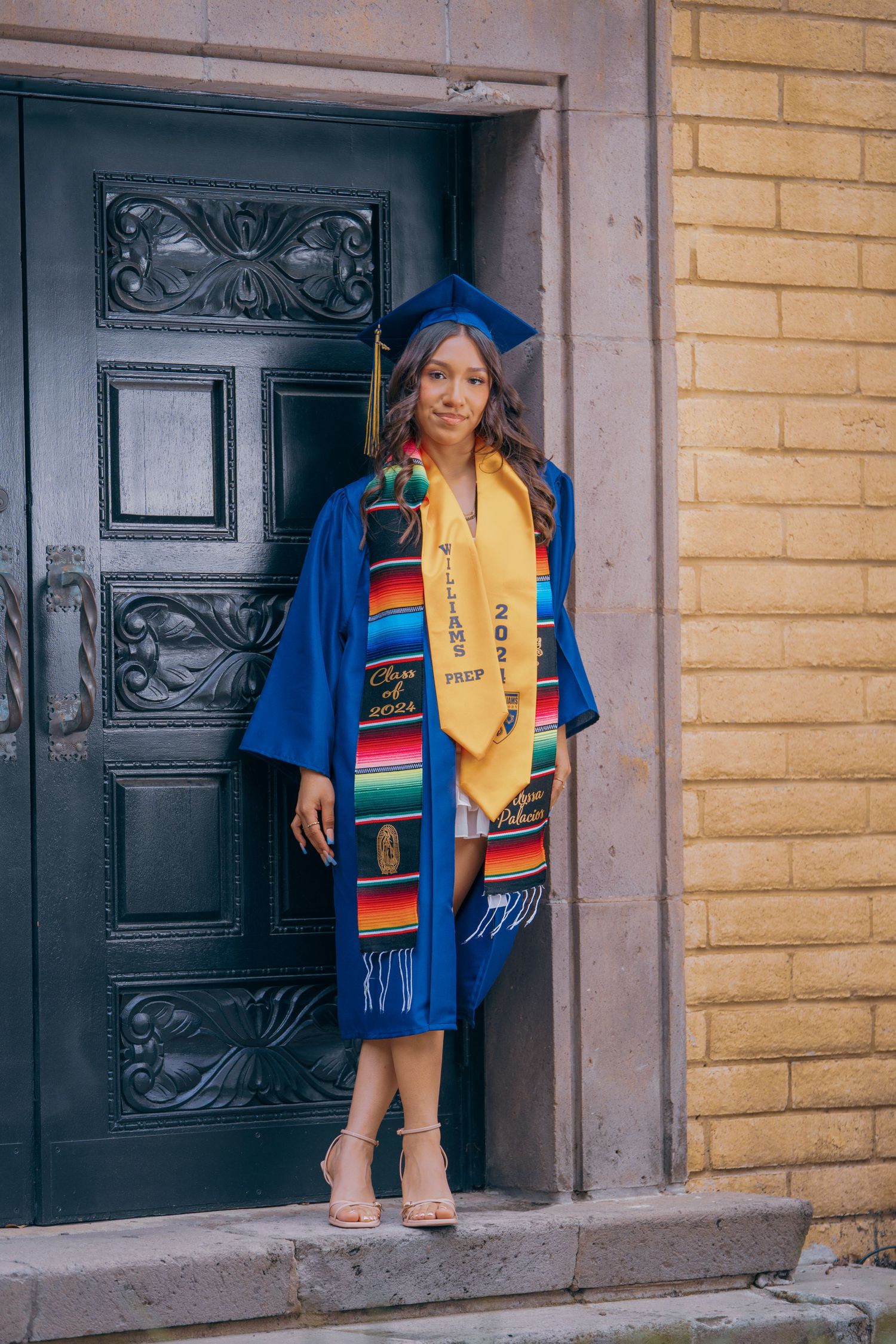 Graduate wearing blue academic regalia and rainbow serape stole stands in front of yellow brick building.