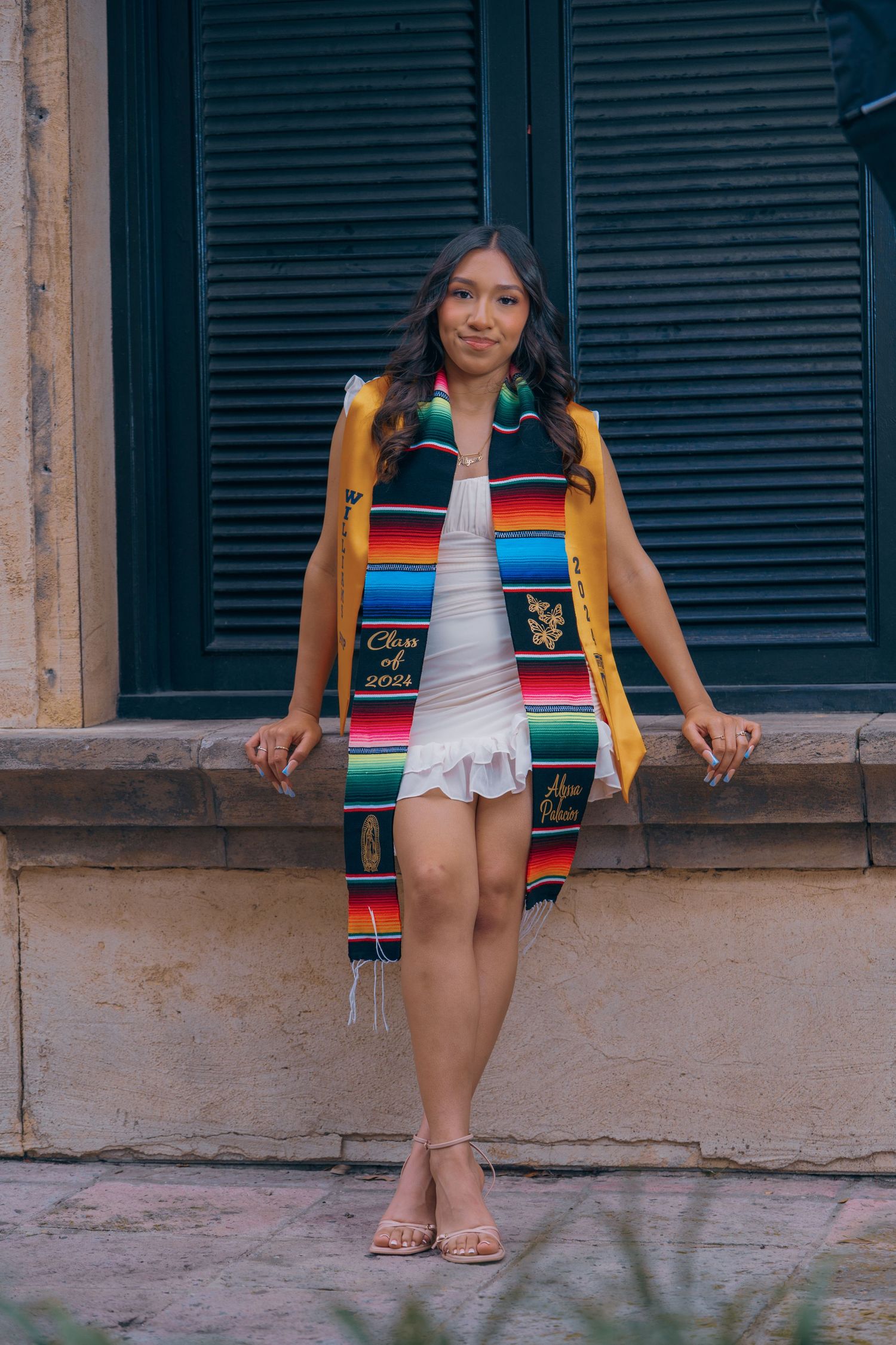 Woman wearing a colorful serape-style top and white shorts sits casually on a concrete ledge outdoors.