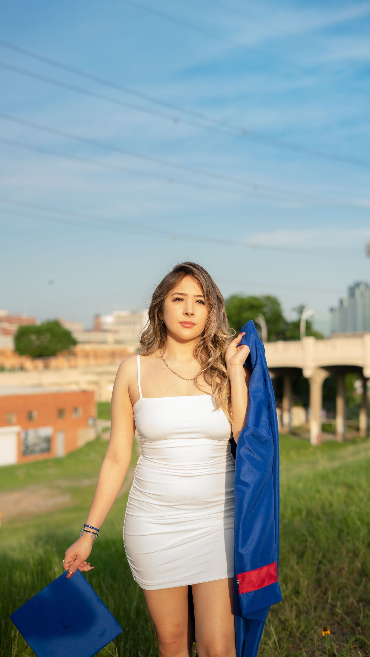 A person in a white dress stands outdoors holding a blue and red garment on a sunny day with buildings in the background.
