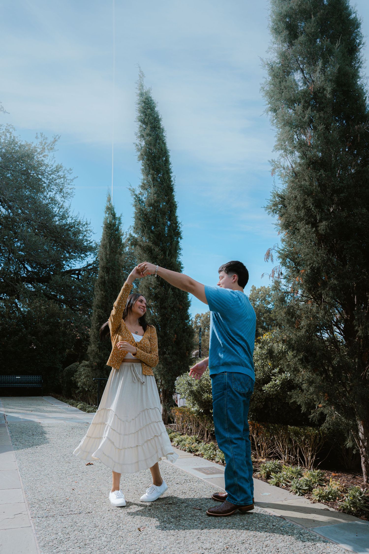 A couple dances together on a garden path surrounded by tall cypress trees on a sunny day.