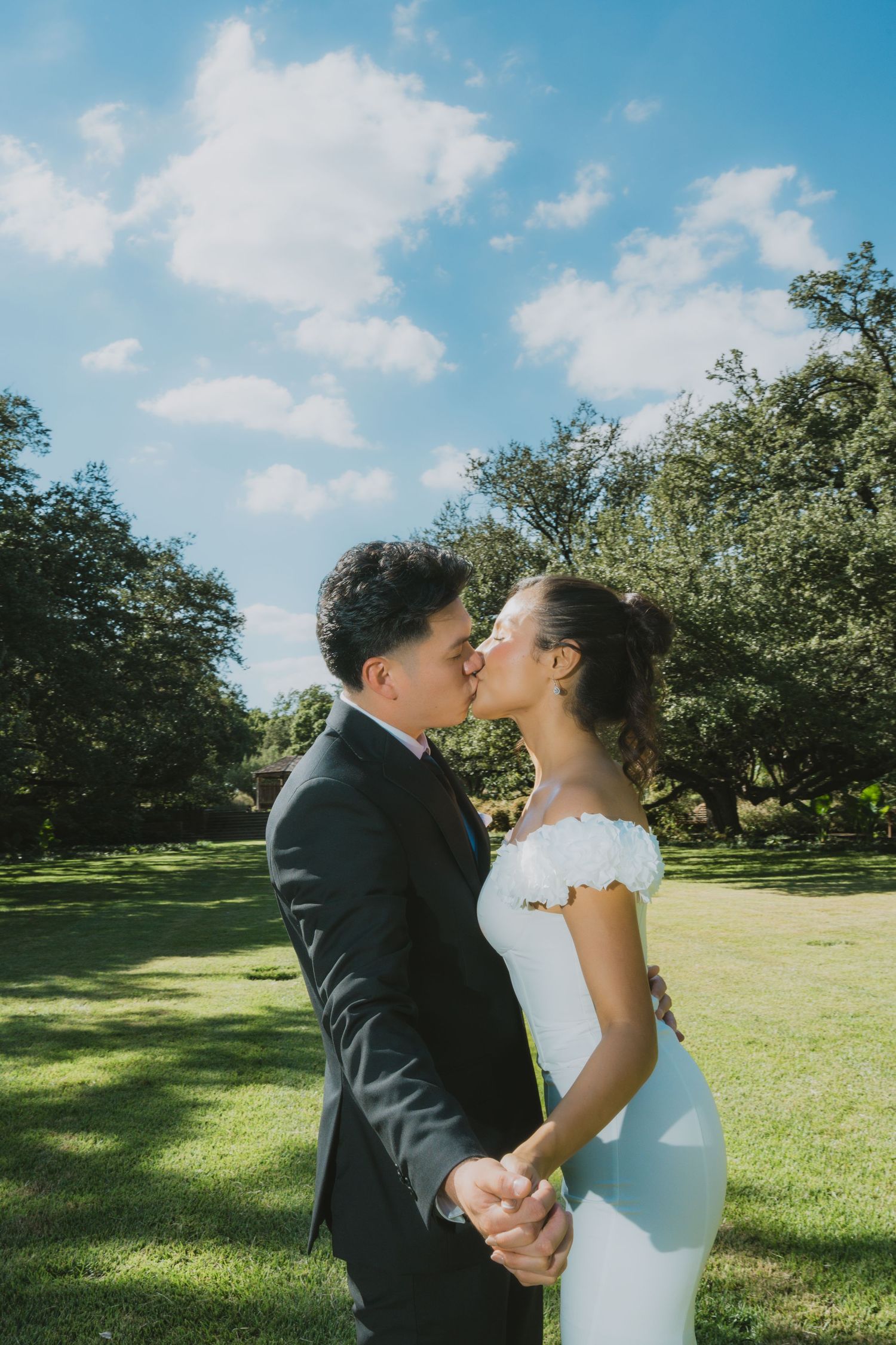 A bride and groom share a romantic kiss outdoors on a sunny day, surrounded by lush green trees and grass.