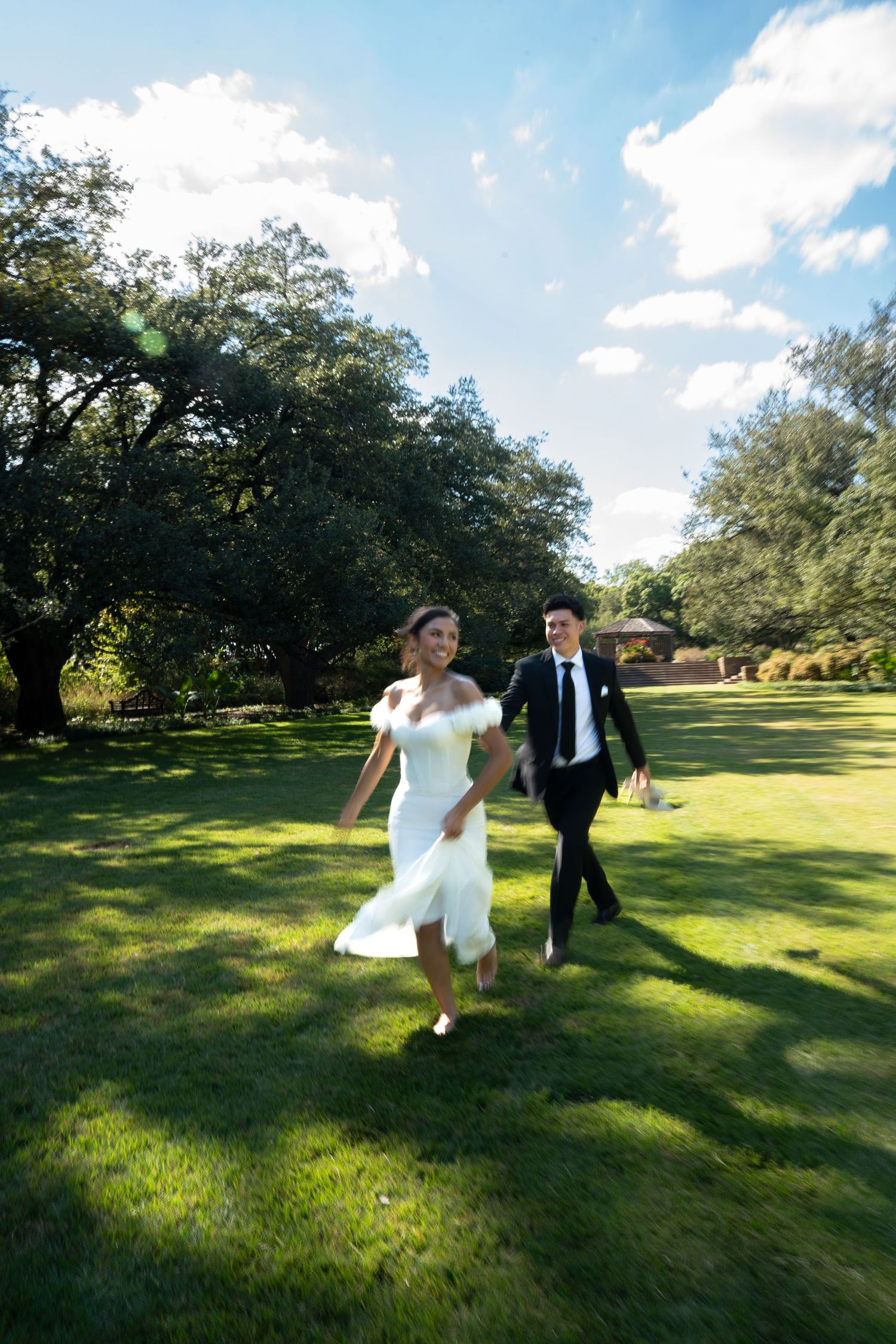 A joyful bride in a white dress and groom in a black suit run hand-in-hand across a lush green lawn.