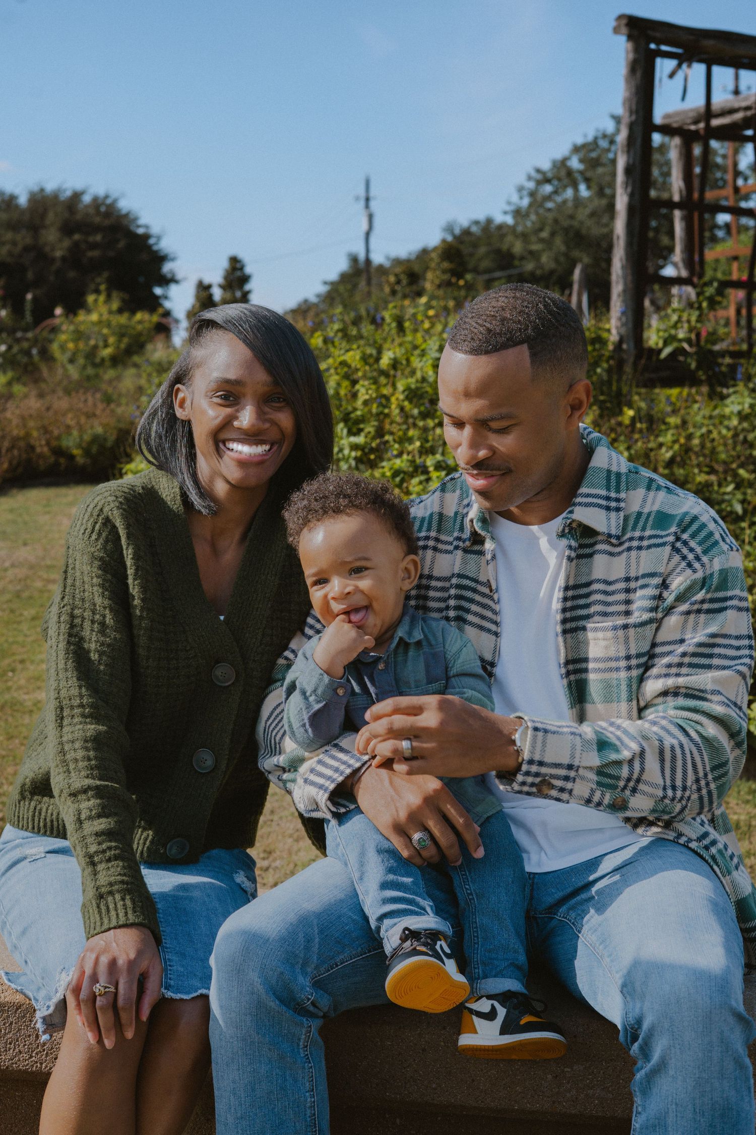 Happy Black family with parents and toddler son sitting outdoors on sunny day in casual denim outfits.