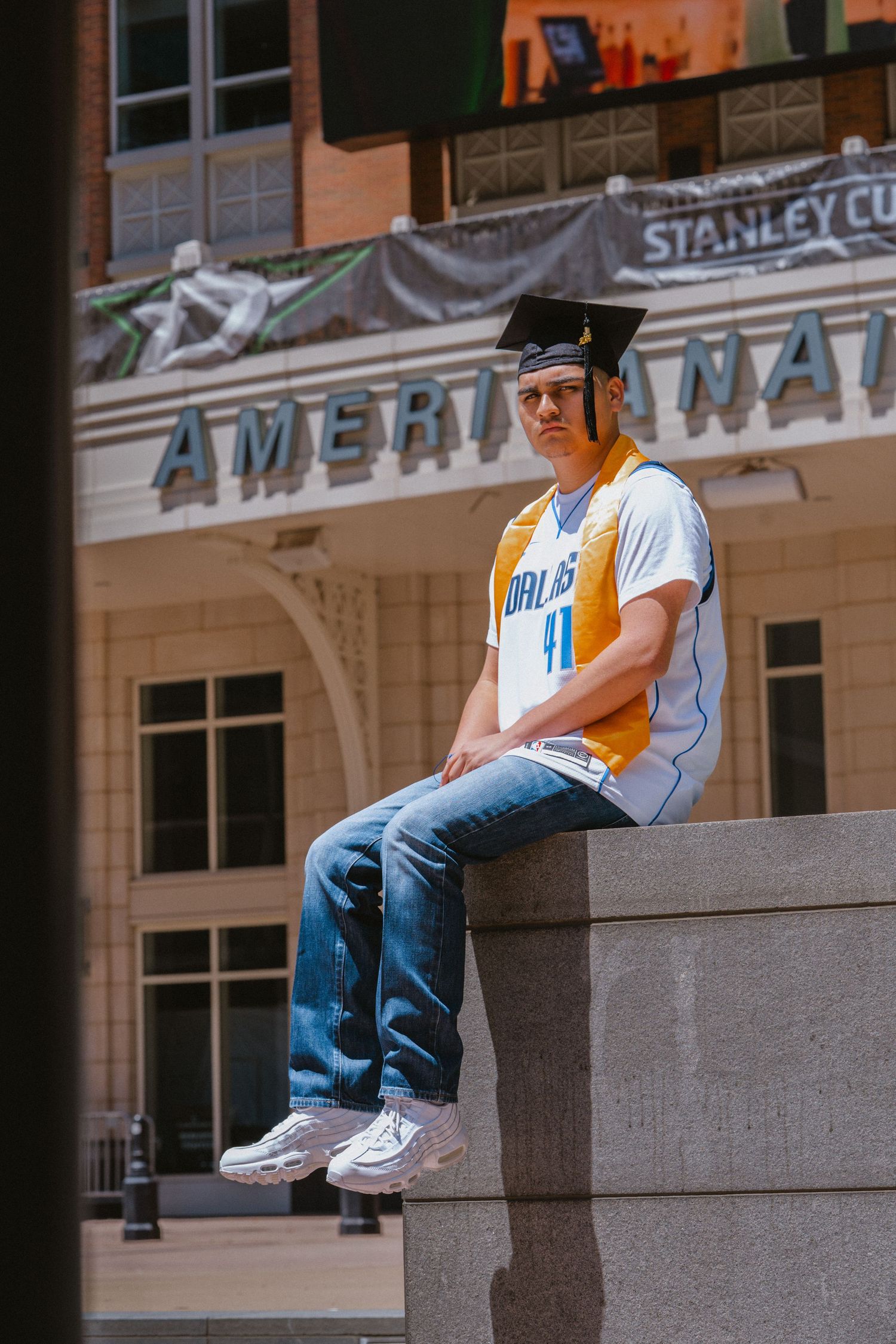 Graduate in cap and Mavericks jersey sits on concrete ledge outside American Airlines Center in Dallas.