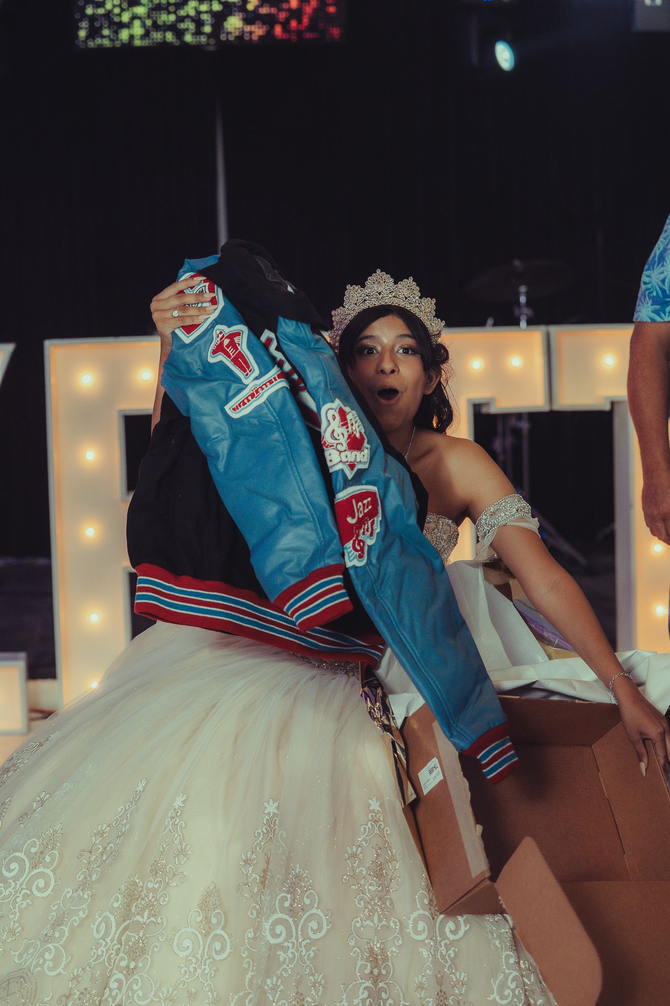 Young woman in a tiara and white ballgown hugging a blue varsity jacket at a quinceañera celebration.