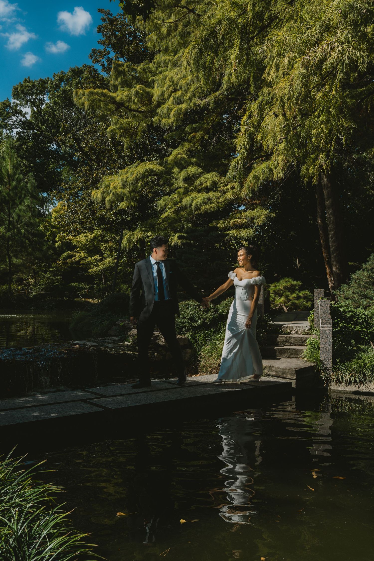 Bride and groom hold hands walking beside a serene garden pond surrounded by lush green trees on a sunny day.