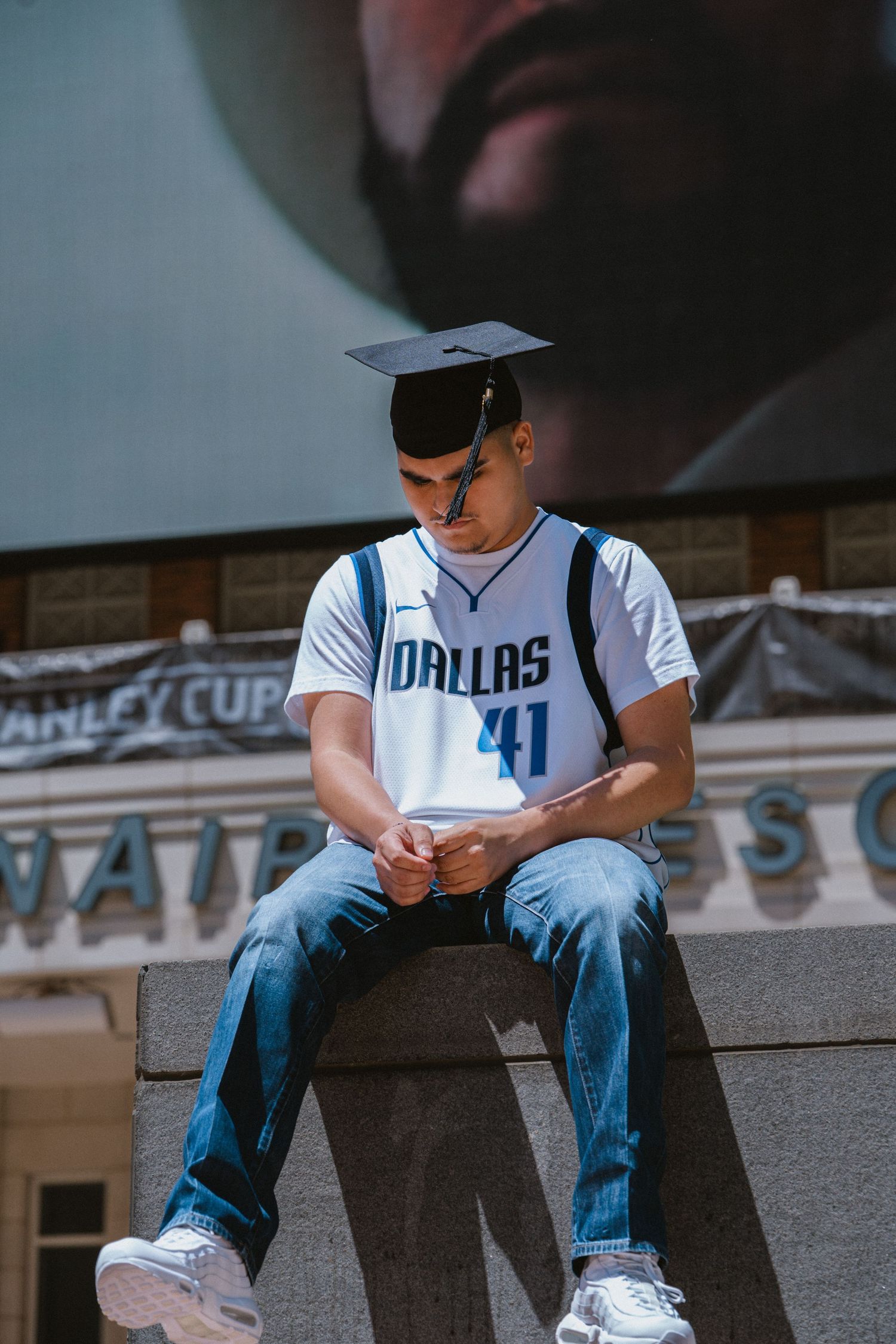 Young man in Dallas 41 jersey and graduation cap sits on stone ledge looking down at his hands outdoors.